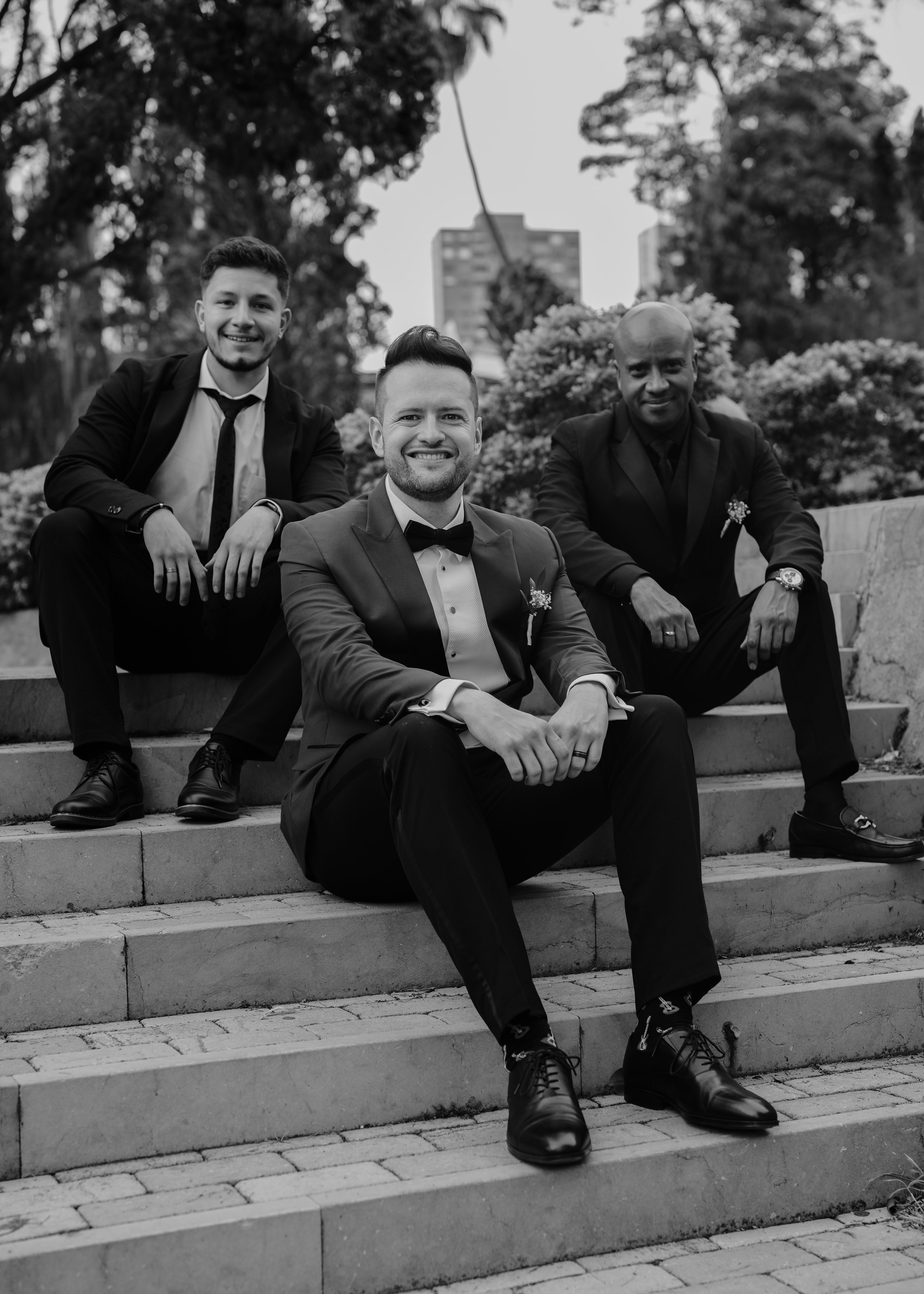 Three men in suits sitting on outdoor steps, smiling, with trees and a high-rise building in the background. Two are wearing tuxedos with bow ties, and one is in a suit with a regular tie.