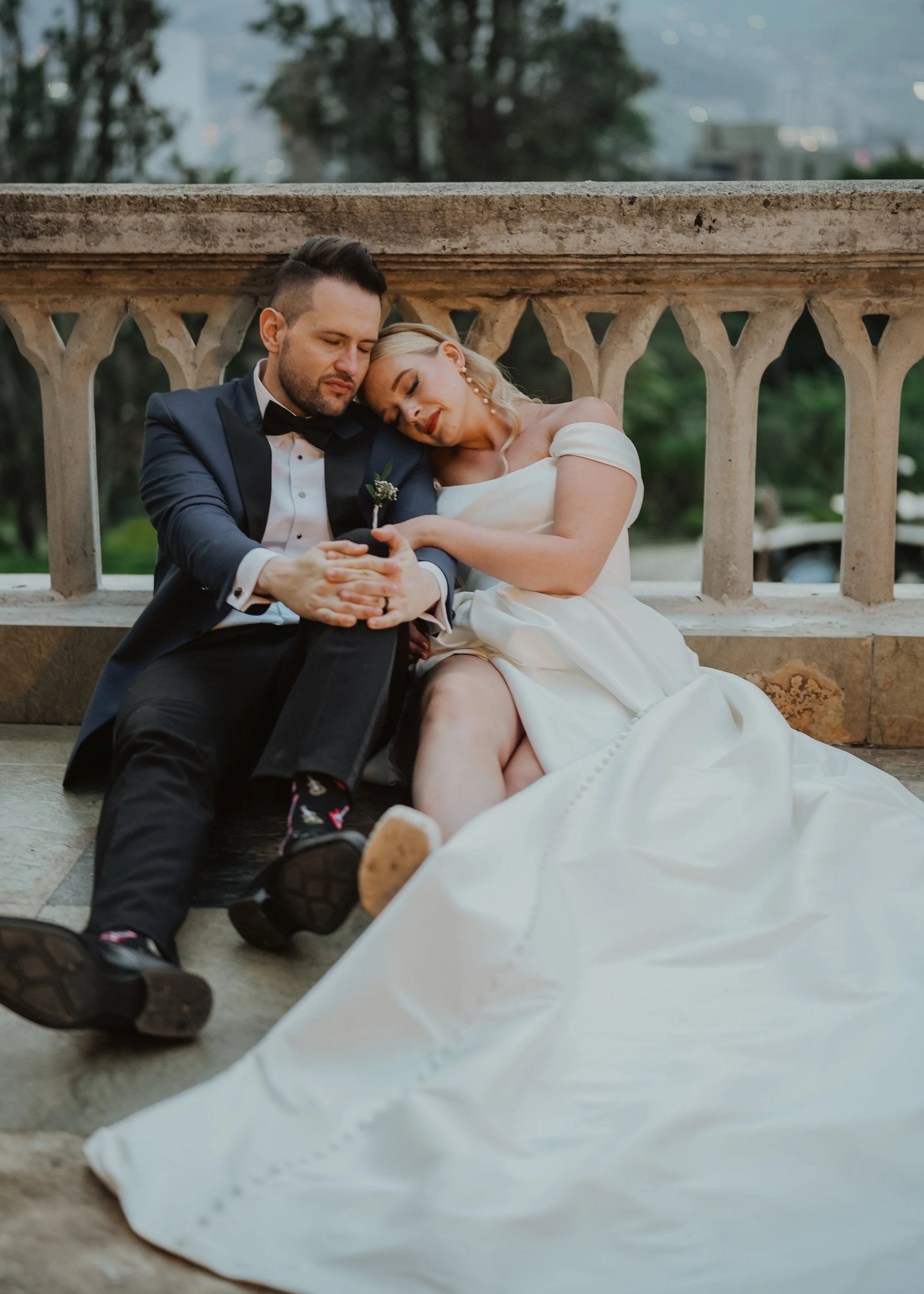A bride and groom sitting on the ground and leaning against a stone railing. The bride's head rests on the groom's shoulder, and they both have their eyes closed in a peaceful moment. The groom is wearing a tuxedo with a bow tie, and the bride is in 