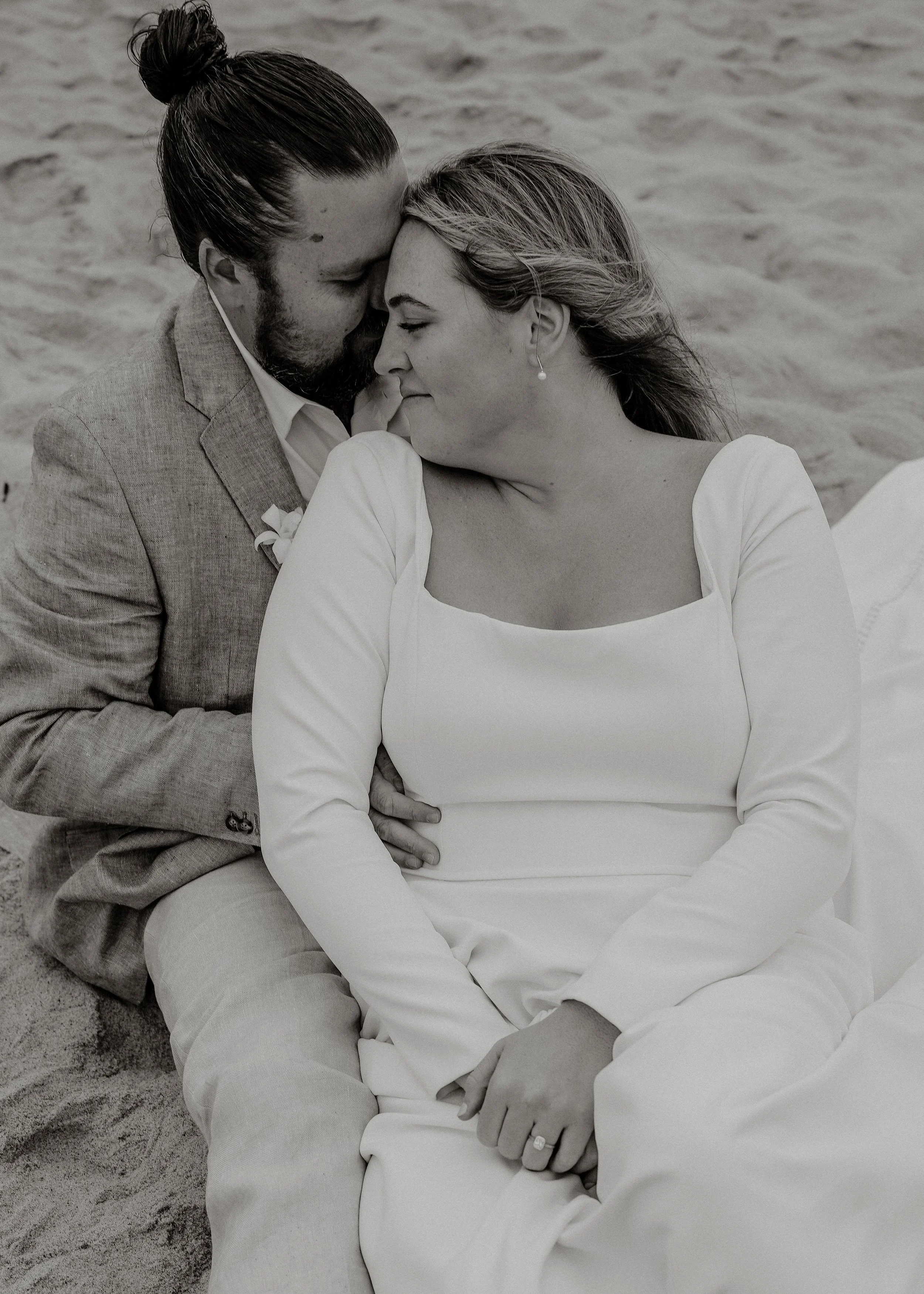 A black-and-white photo of a couple embracing and touching foreheads on a sandy surface, with the woman wearing a white top and the man wearing a tan blazer.