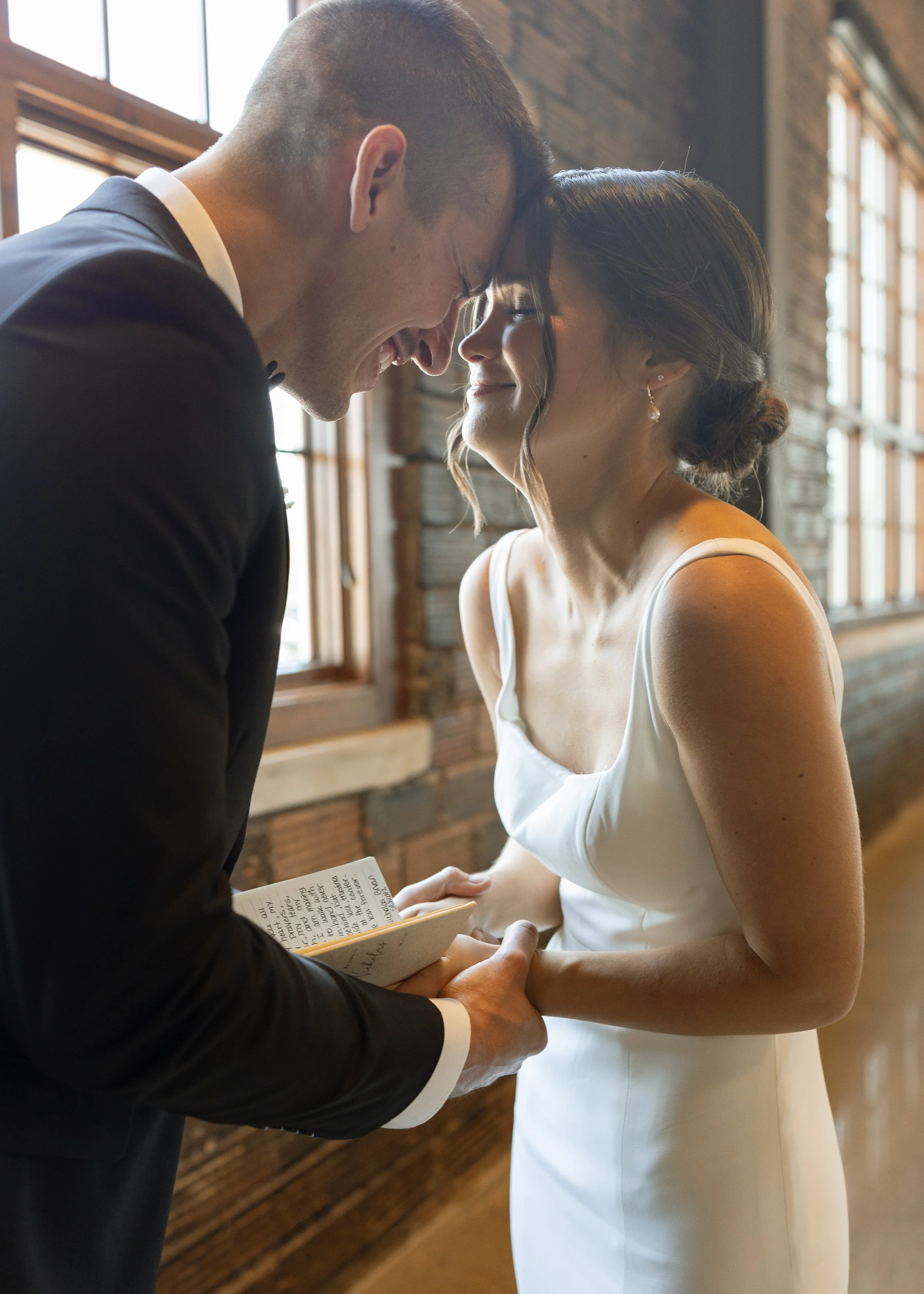 A bride and groom sharing a forehead touch during their wedding ceremony, both smiling gently with their eyes closed, indoors in front of a large window with wooden framing.