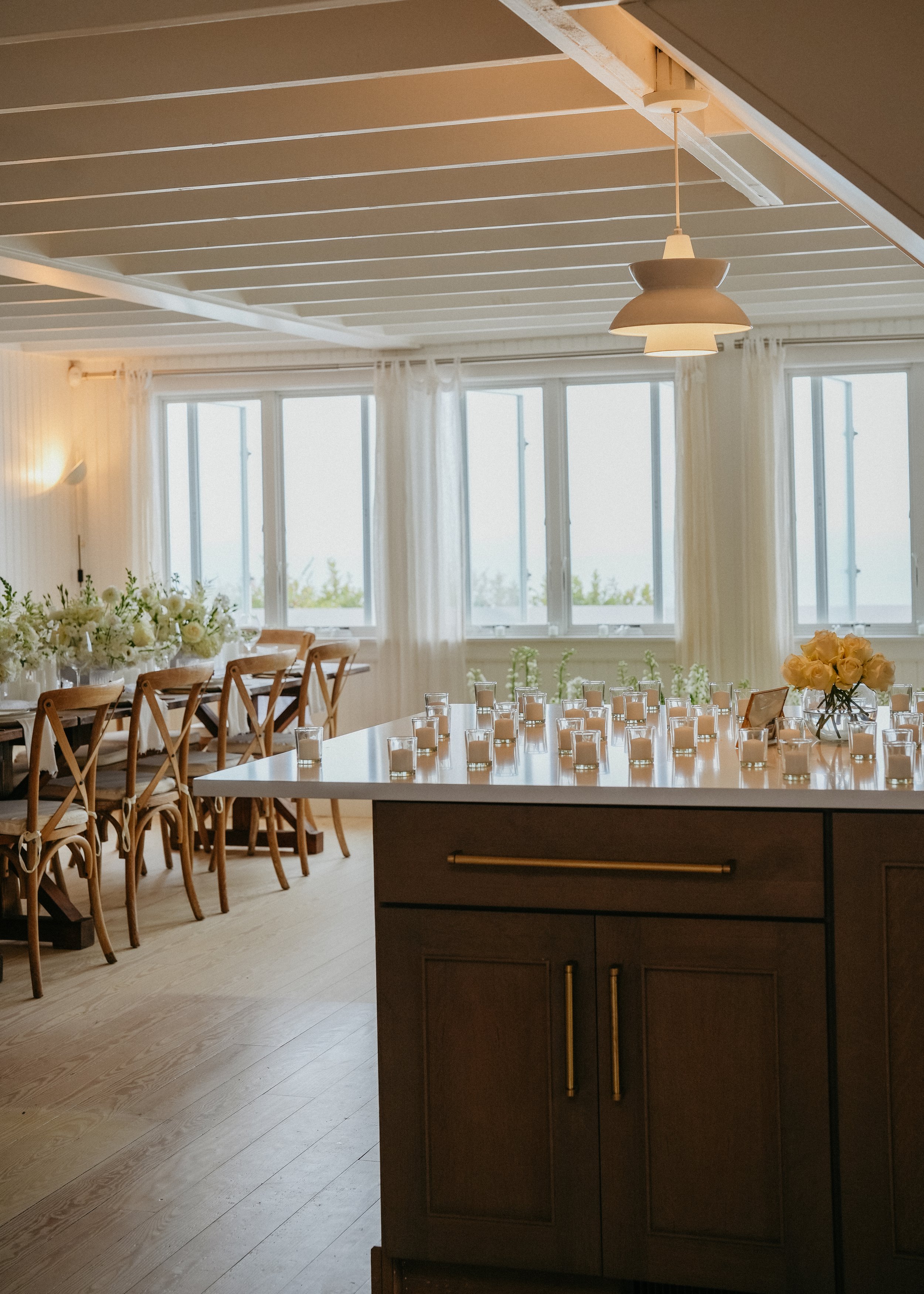 A dining room with a view of large windows, decorated with flowers and candle holders on a table, and a modern hanging light fixture.