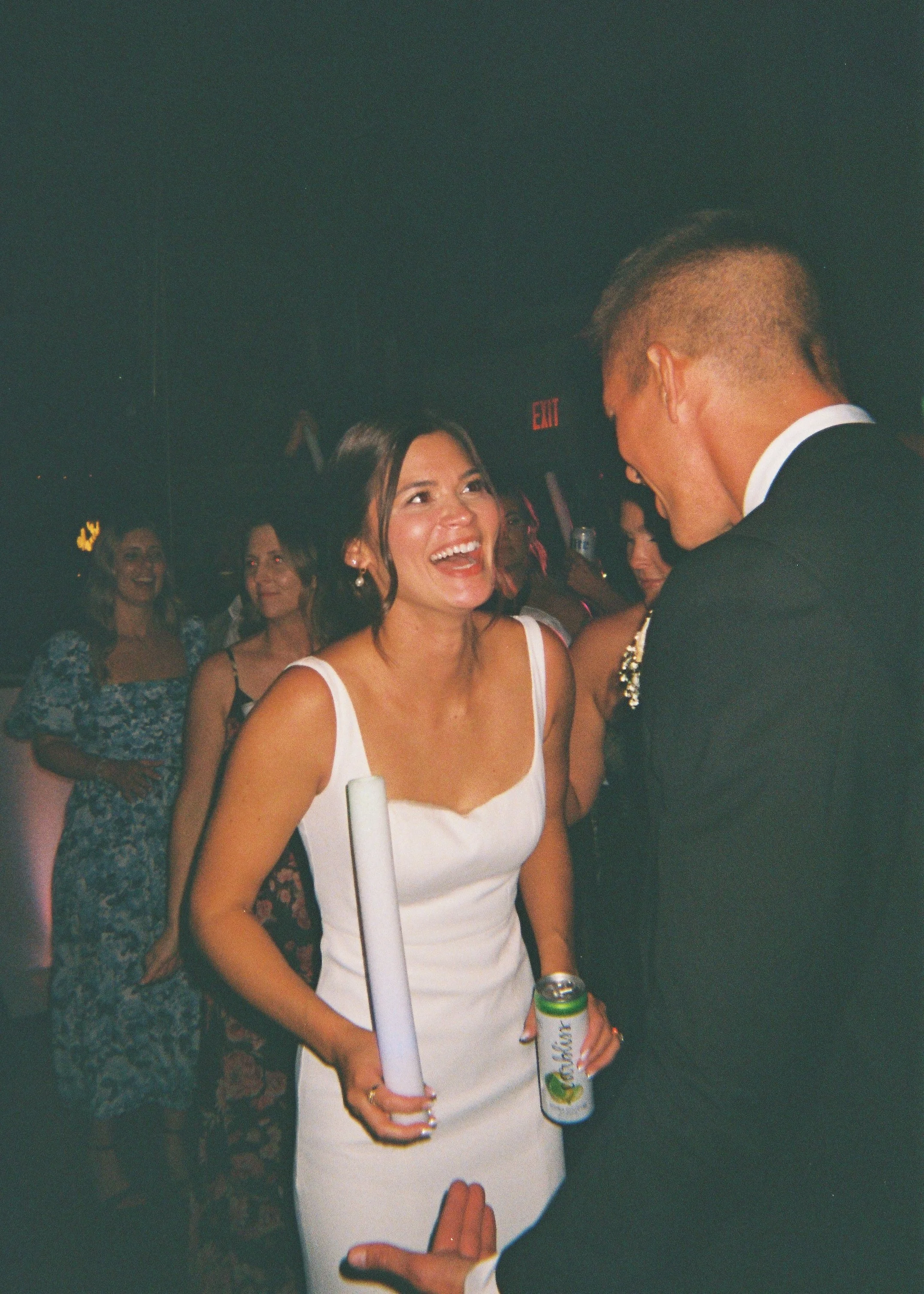A woman in a white dress laughing with a man in a suit at a social gathering, multiple women in the background, dim lighting, and an 'EXIT' sign visible.
