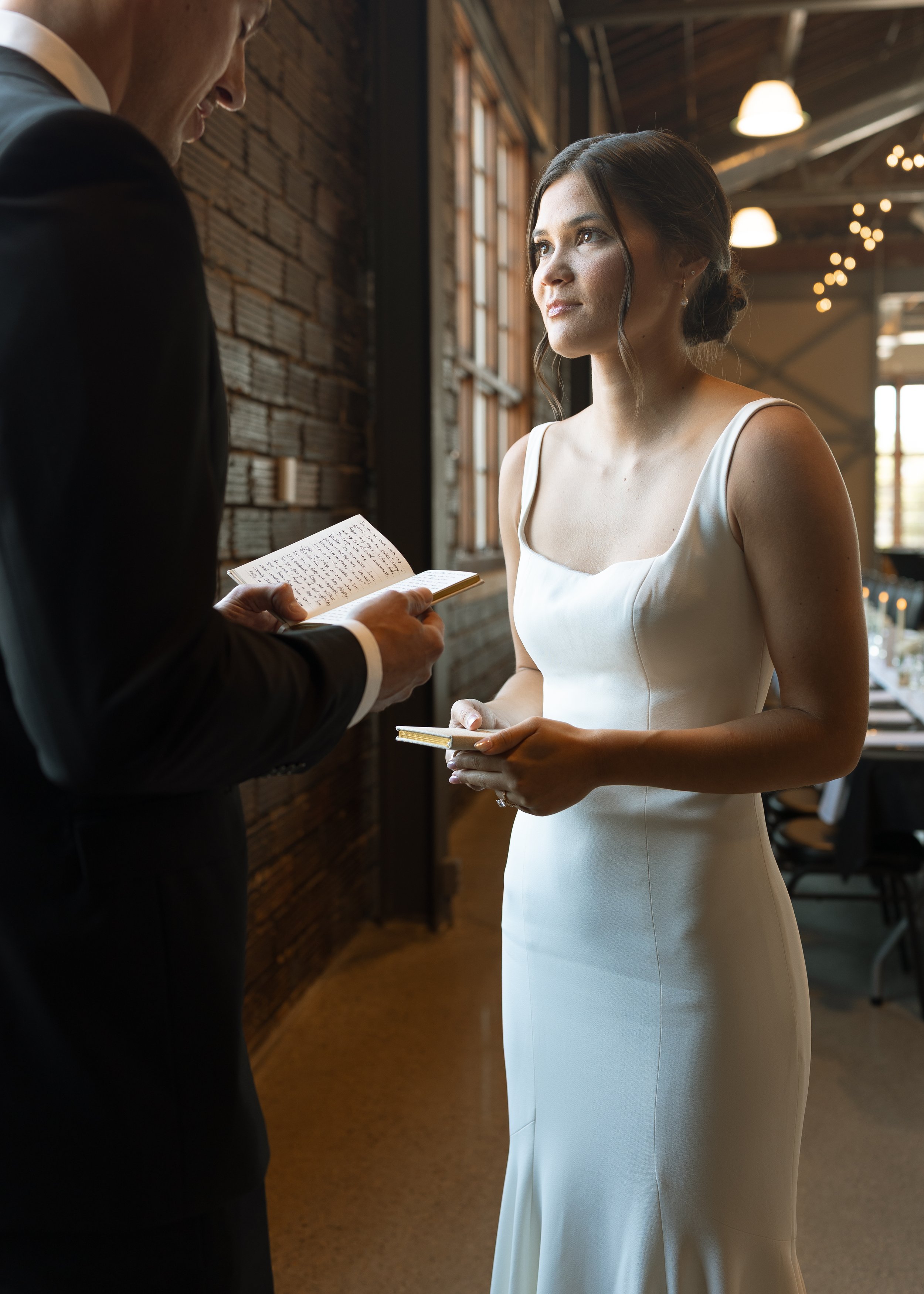 A woman in a white wedding dress standing in front of a man in a formal suit, exchanging vows during a wedding ceremony inside a rustic venue with exposed brick walls and large windows.