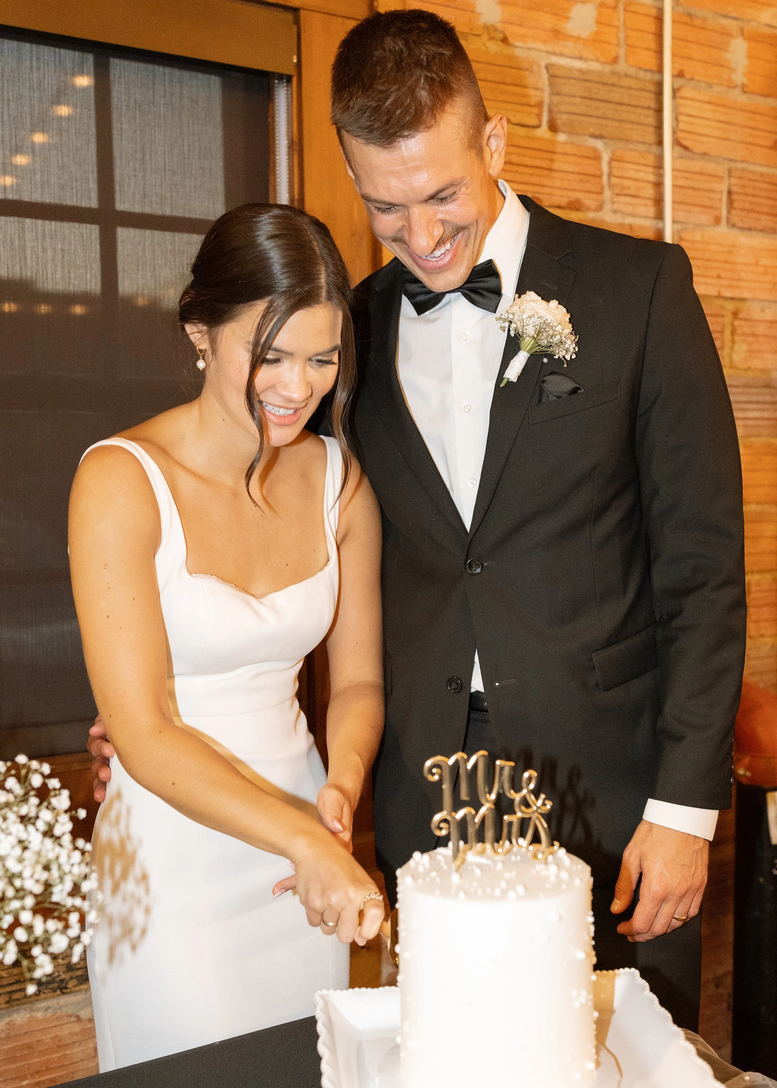 A bride and groom are cutting a wedding cake together. The bride is wearing a white dress, and the groom is wearing a black tuxedo with a bow tie and a boutonniere. They are smiling and looking down at the cake.