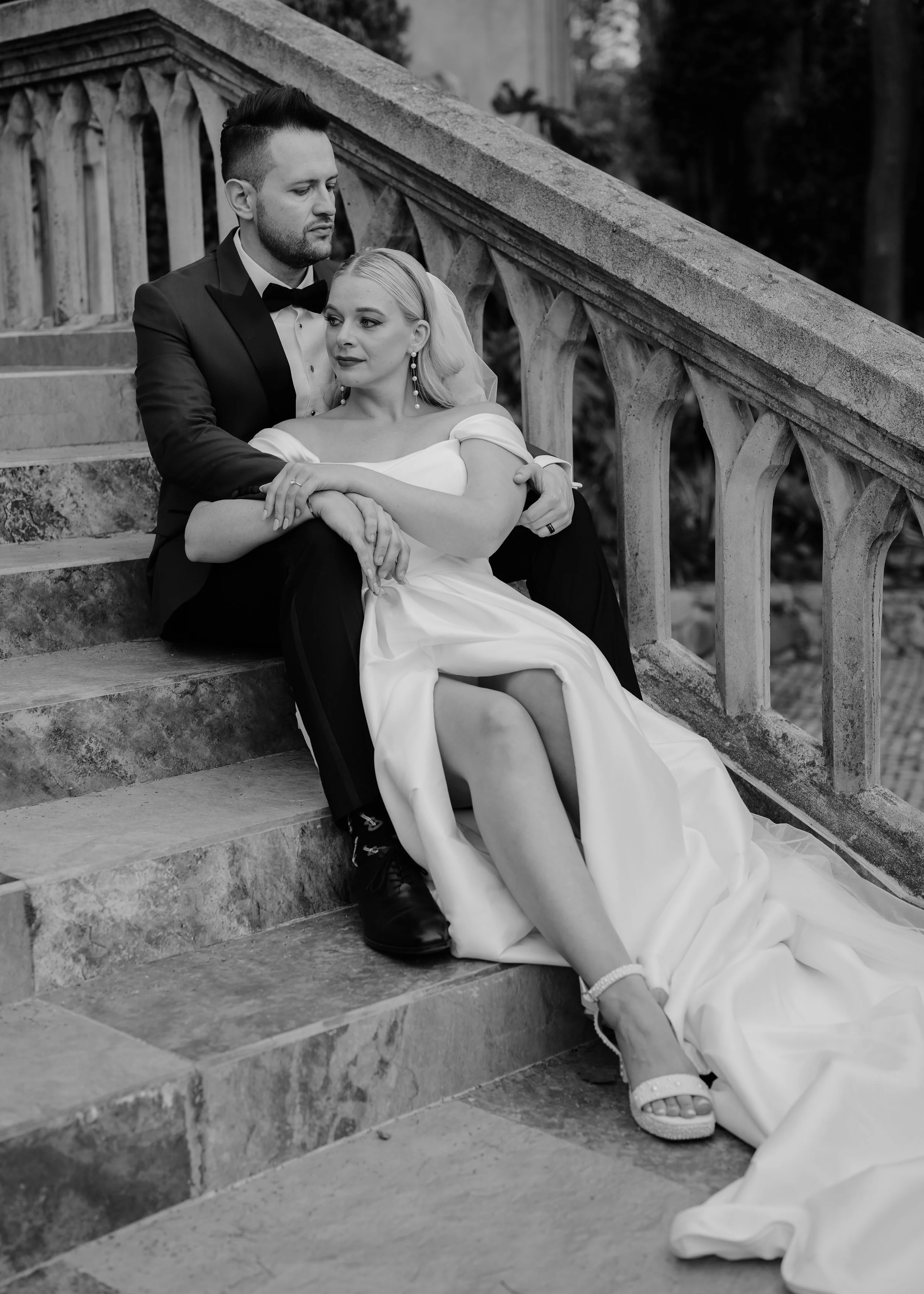 Black and white photograph of a bride and groom sitting on outdoor stone stairs. The groom in a tuxedo is sitting behind the bride, holding her shoulders, as she leans against him with a calm expression. The bride is wearing a strapless wedding gown 