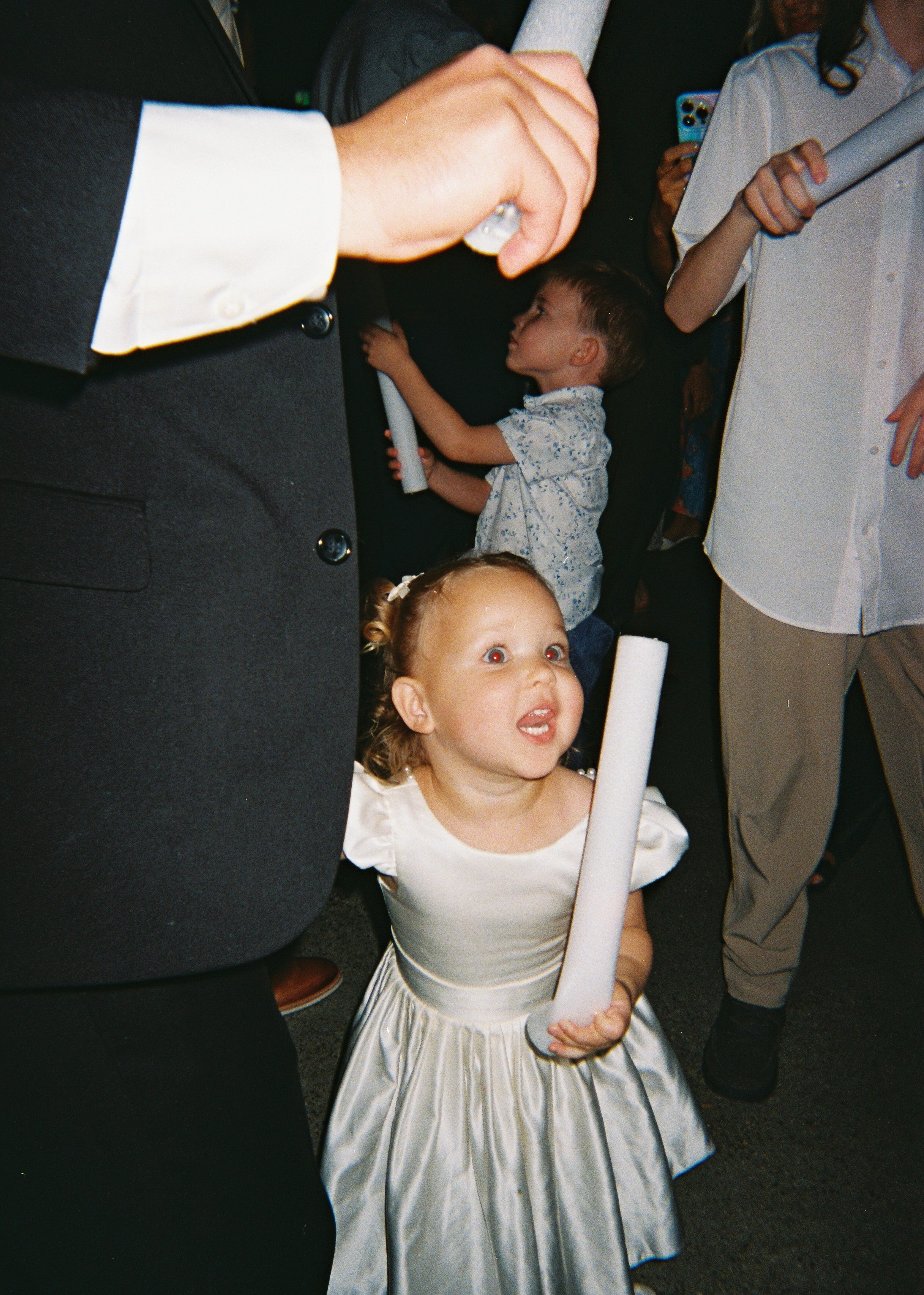 A young girl in a white dress holding a white candle, surrounded by people at a ceremony or celebration.
