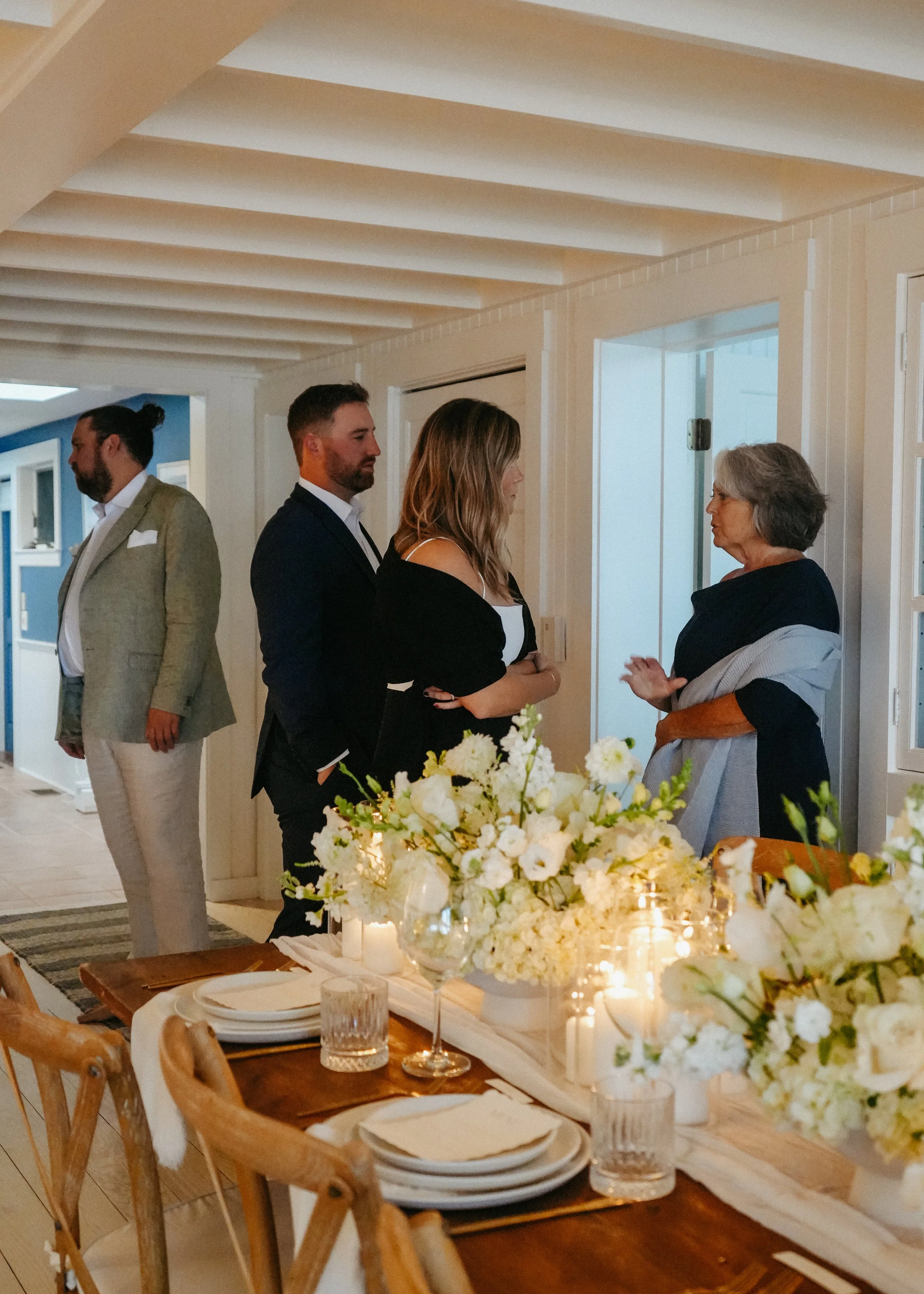 People standing and talking in a well-lit room, with a table set with floral arrangements, candles, glassware, plates, and napkins in the foreground.