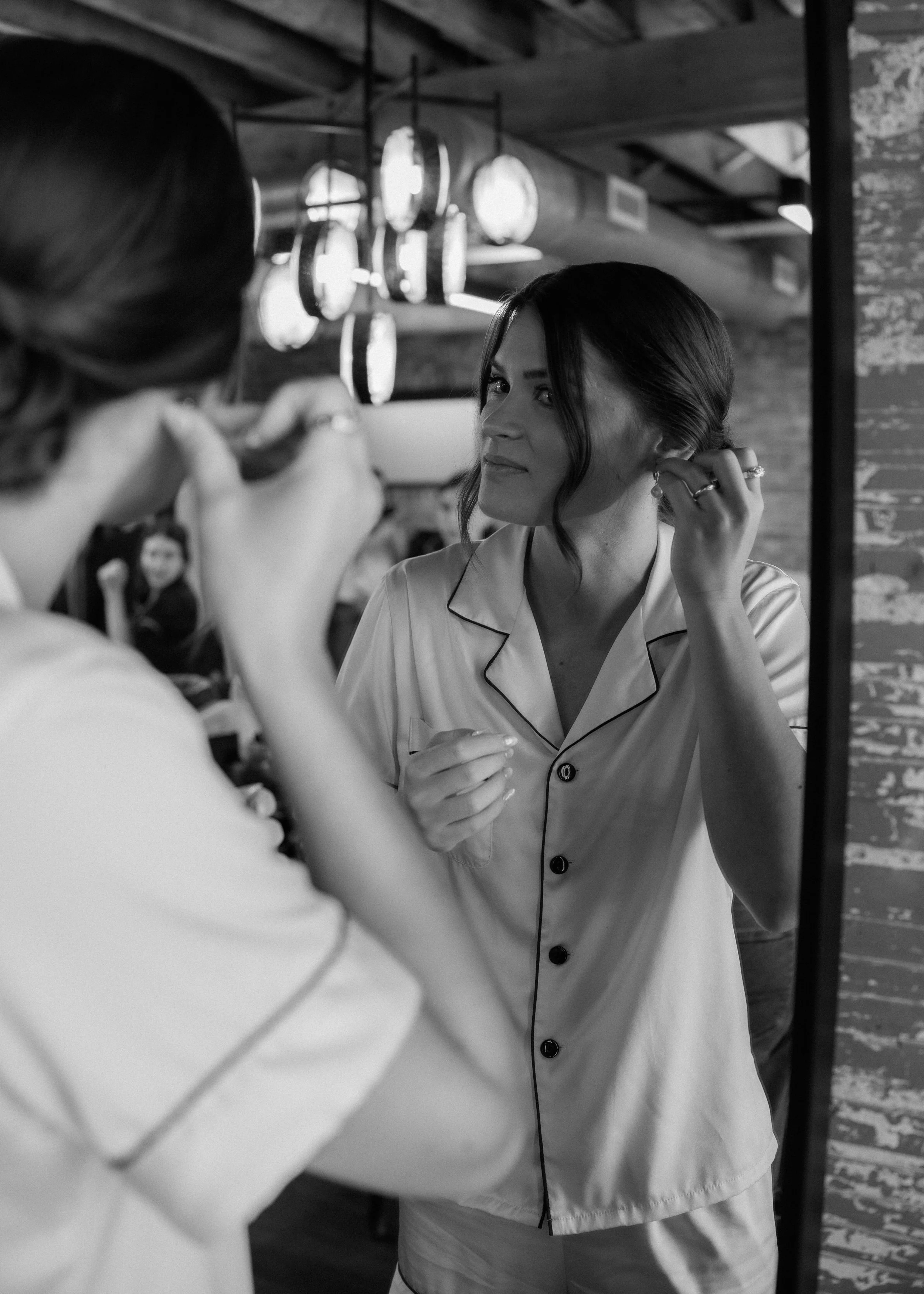 A woman in pajamas is putting on an earring while another person, whose face is not visible, is adjusting her earring in a cozy, rustic room with hanging pots or pans on the wall.