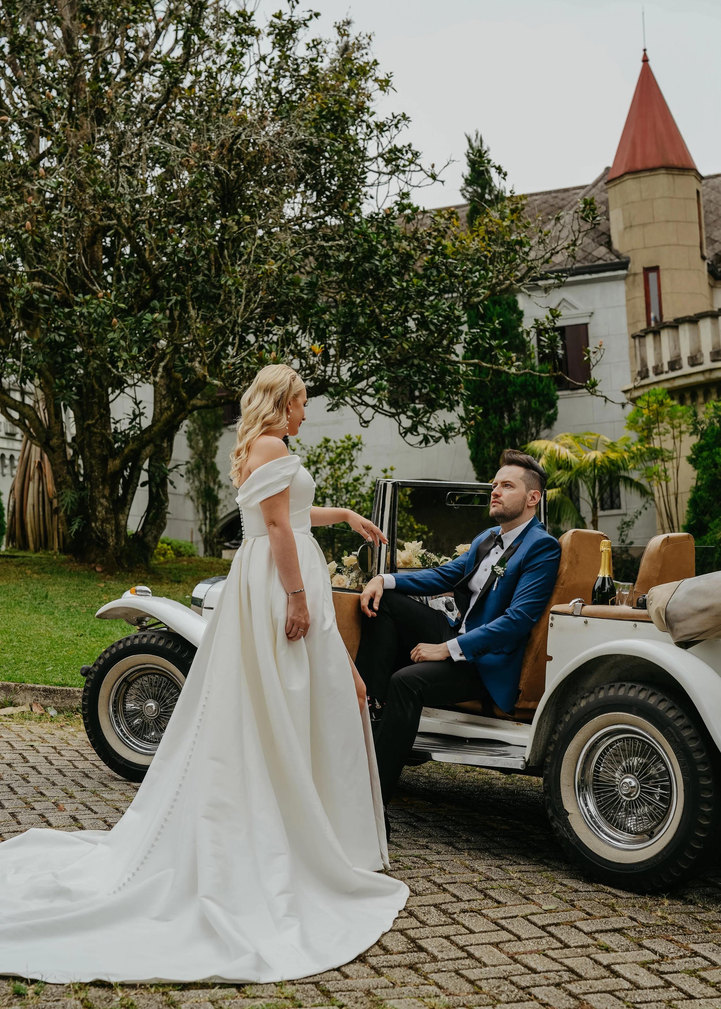 A bride and groom at a wedding, with the bride standing outside a vintage car and the groom sitting inside, in front of a large house with trees and greenery.