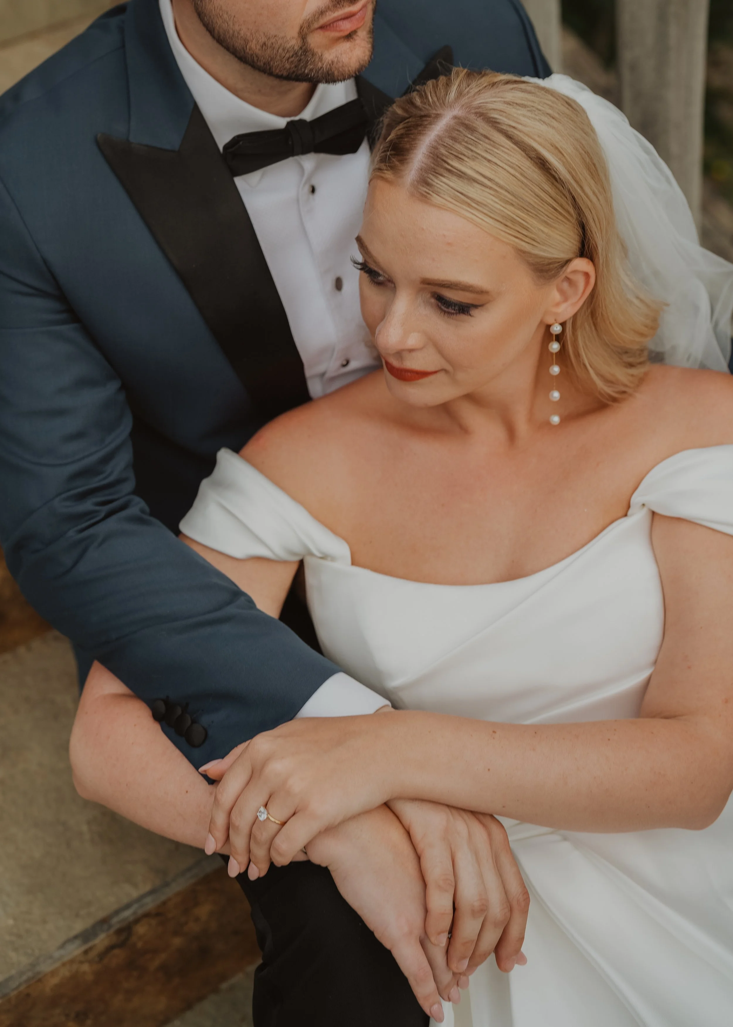 Close-up of a bride and groom embracing, focusing on their hands and part of their faces. The groom wears a tuxedo with a black bow tie, and the bride has blonde hair, earrings, and a white dress.