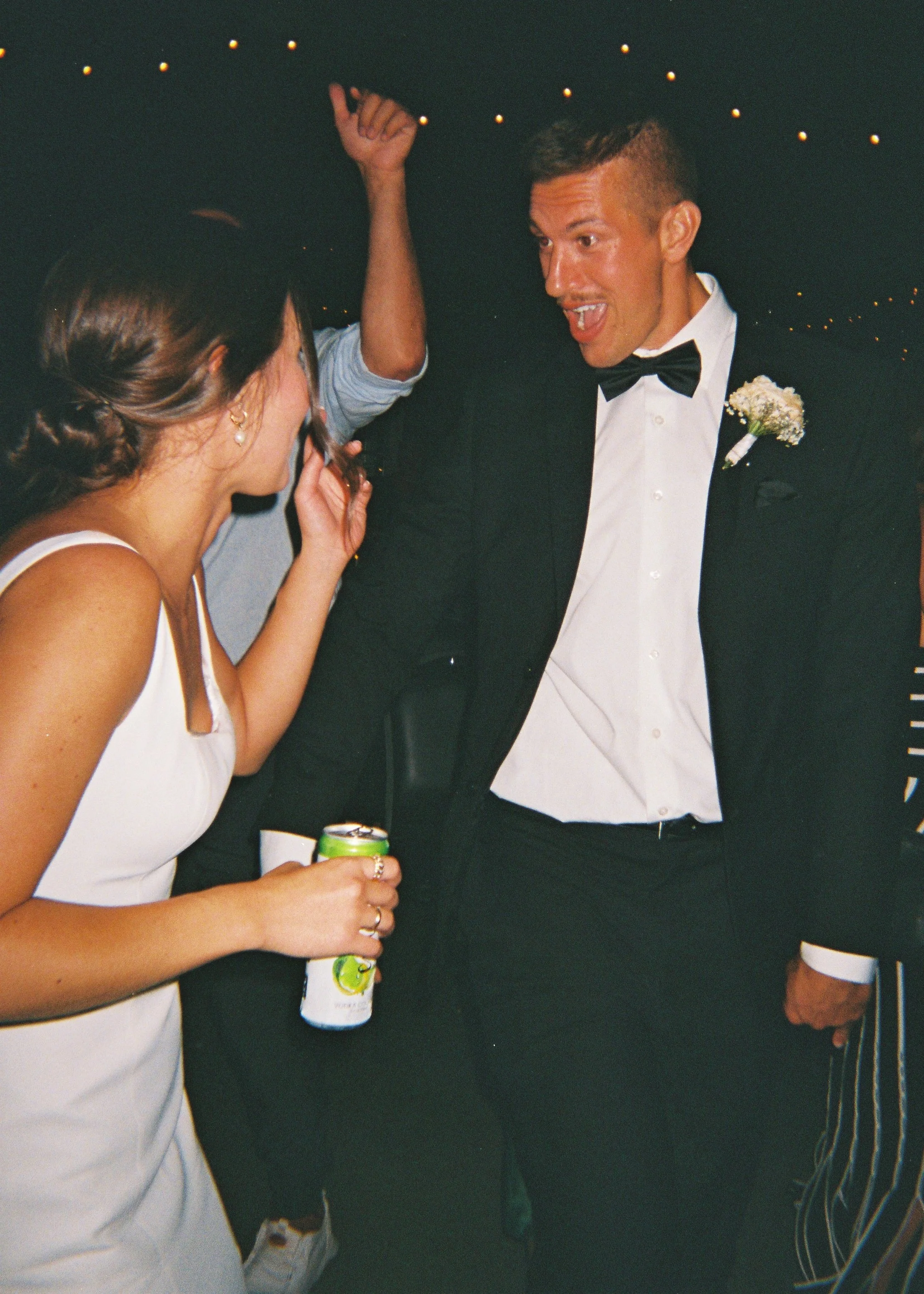 People at a social gathering or party, with a man in a tuxedo and a woman in a white dress, drinking a beverage and engaging in conversation.
