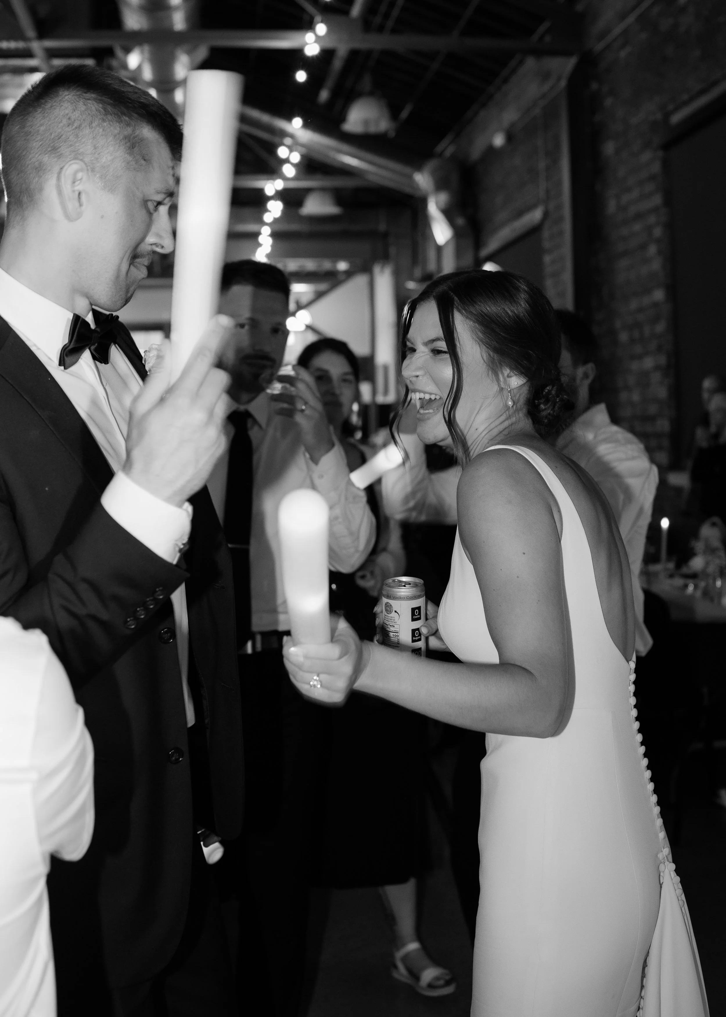 A bride in a white wedding dress laughing and holding a candle at a wedding reception. A groom in a tuxedo is standing next to her, partially visible, and guests are in the background.