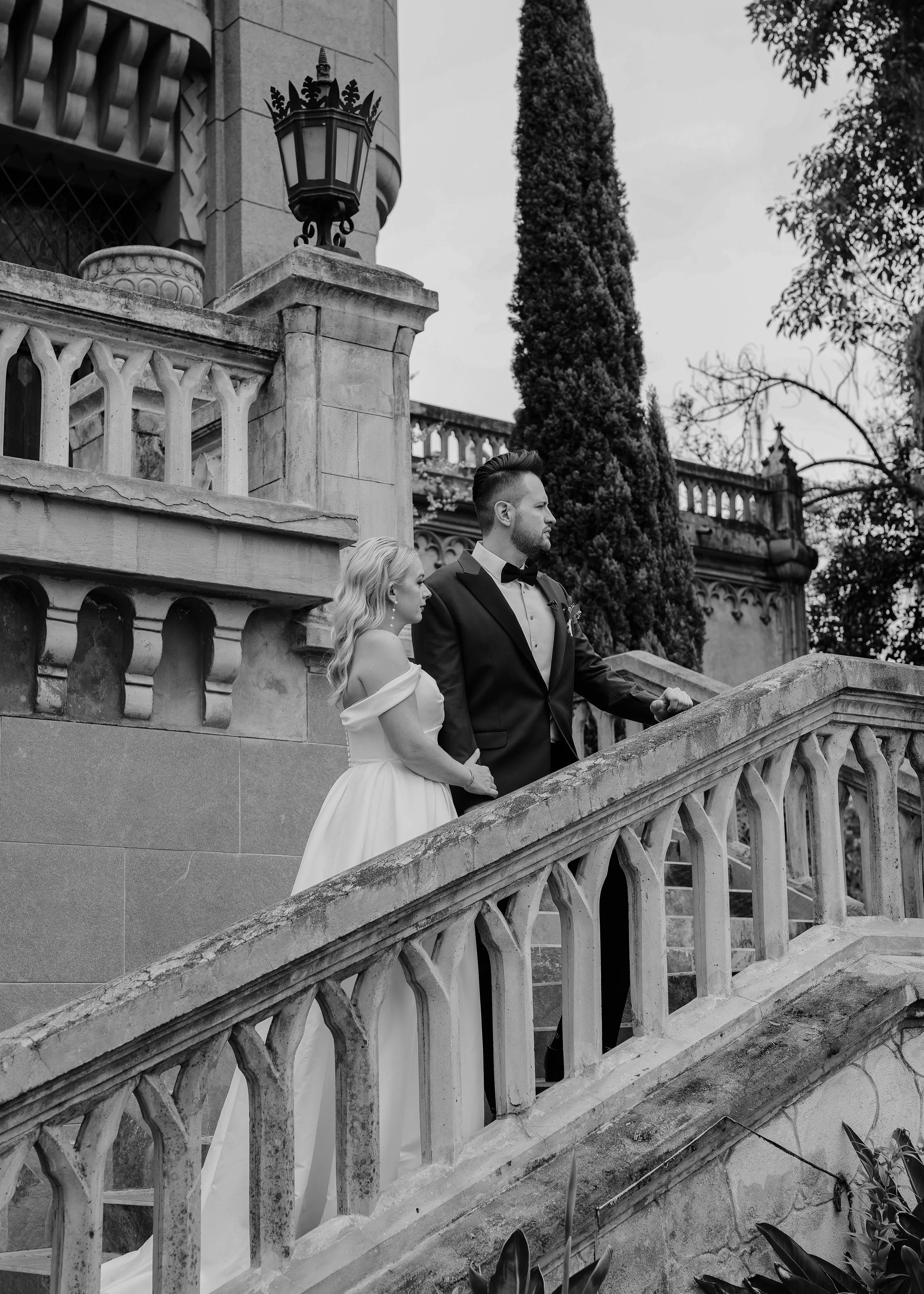 A black-and-white photo of a bride and groom standing on a stone staircase outside a historic building. The bride has long, wavy hair and is wearing an off-shoulder wedding gown. The groom has a beard and hairstyle, dressed in a tuxedo with a bow tie