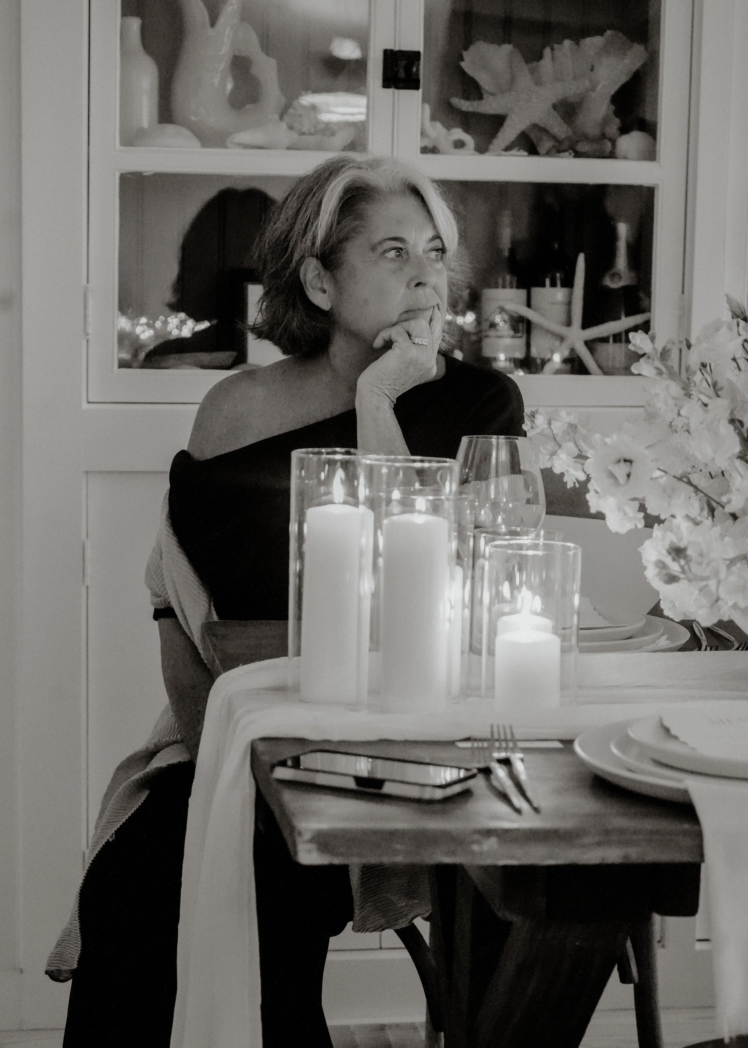 A woman with short, wavy hair with gray streaks is sitting at a table, resting her chin on her hand, and looking thoughtfully to her left. The table is set with lit candles and flowers, and there are plates, cutlery, and glasses in the foreground. Th