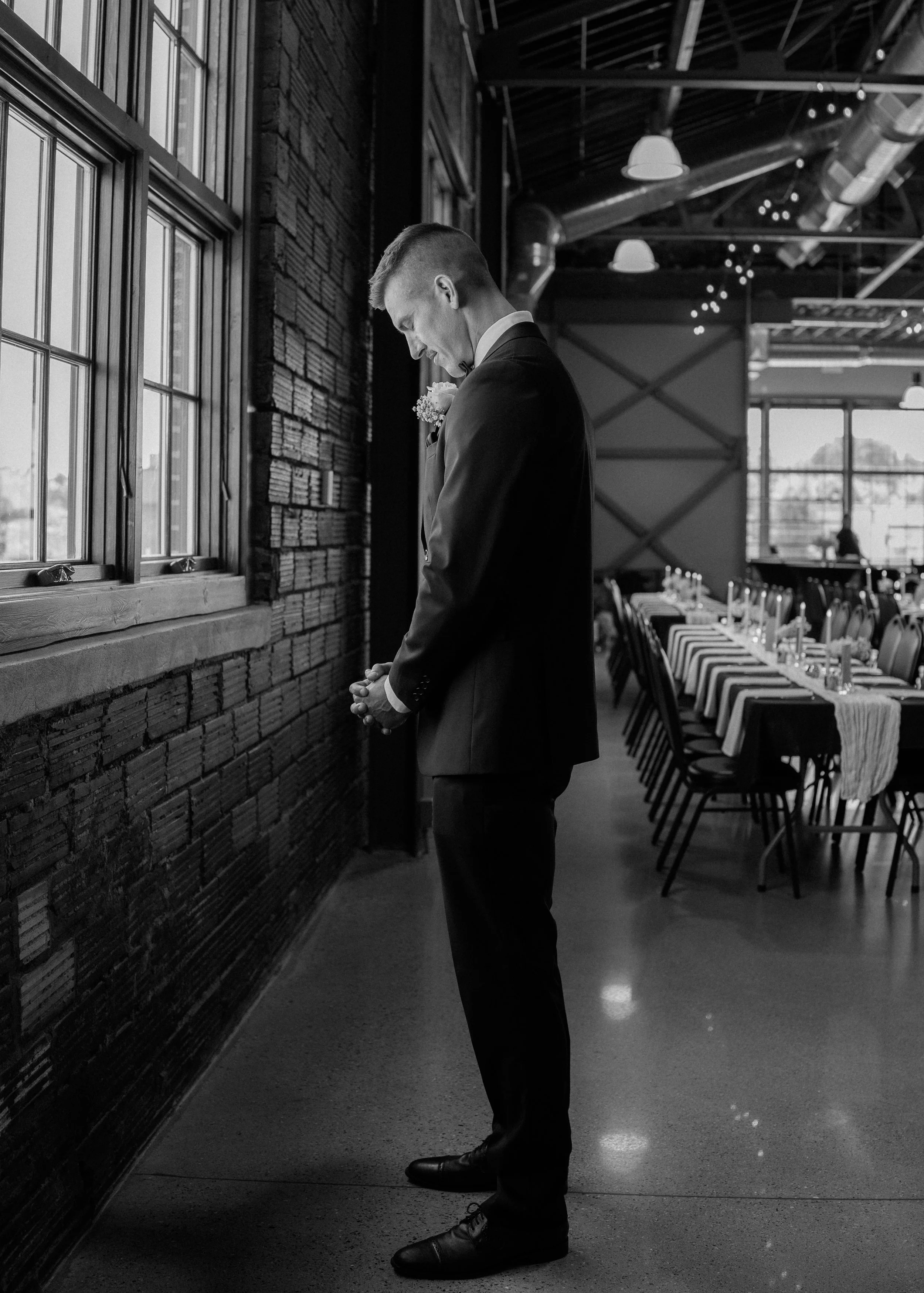 A man in a suit with a boutonniere, likely a groom, standing with his head bowed and hands clasped in front of him, near a window in a dimly lit venue decorated for a wedding or formal event.