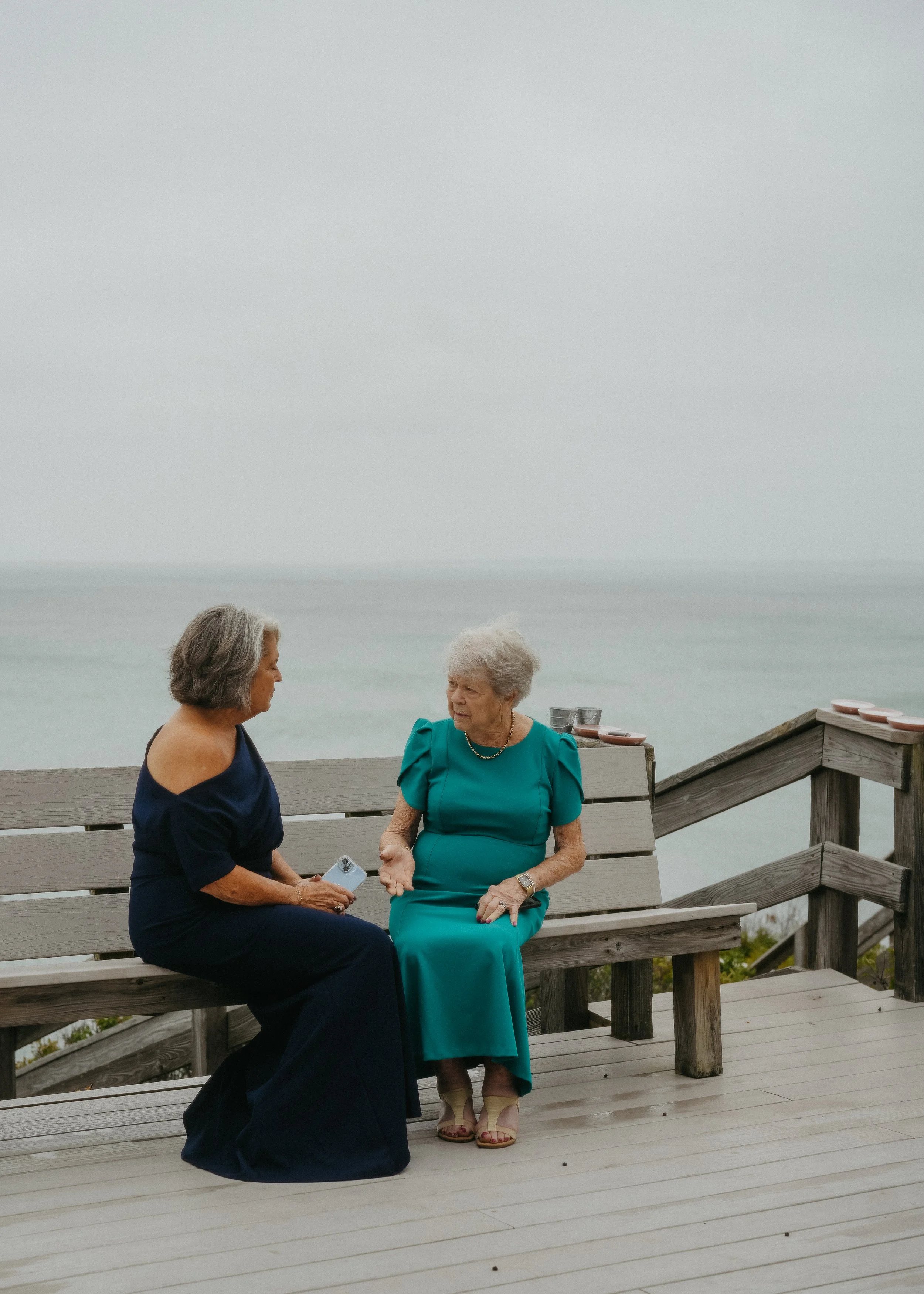 Two women engaged in conversation on a wooden deck overlooking the ocean. One woman is wearing a dark blue off-shoulder dress, and the other is in a teal dress with high heels. There are pink plates and empty glasses on the railing nearby.