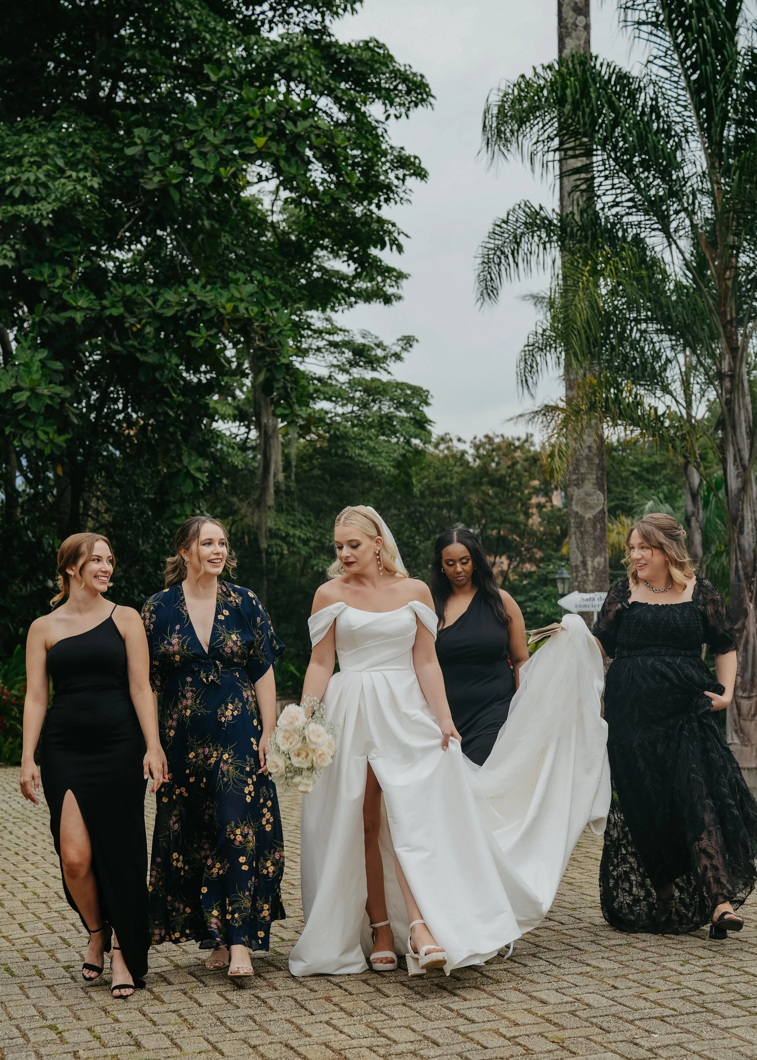 A bride in a white wedding gown with a slit, holding a bouquet of white roses, walking with five women dressed in black or dark-colored dresses in an outdoor garden setting with tall trees and overcast sky.