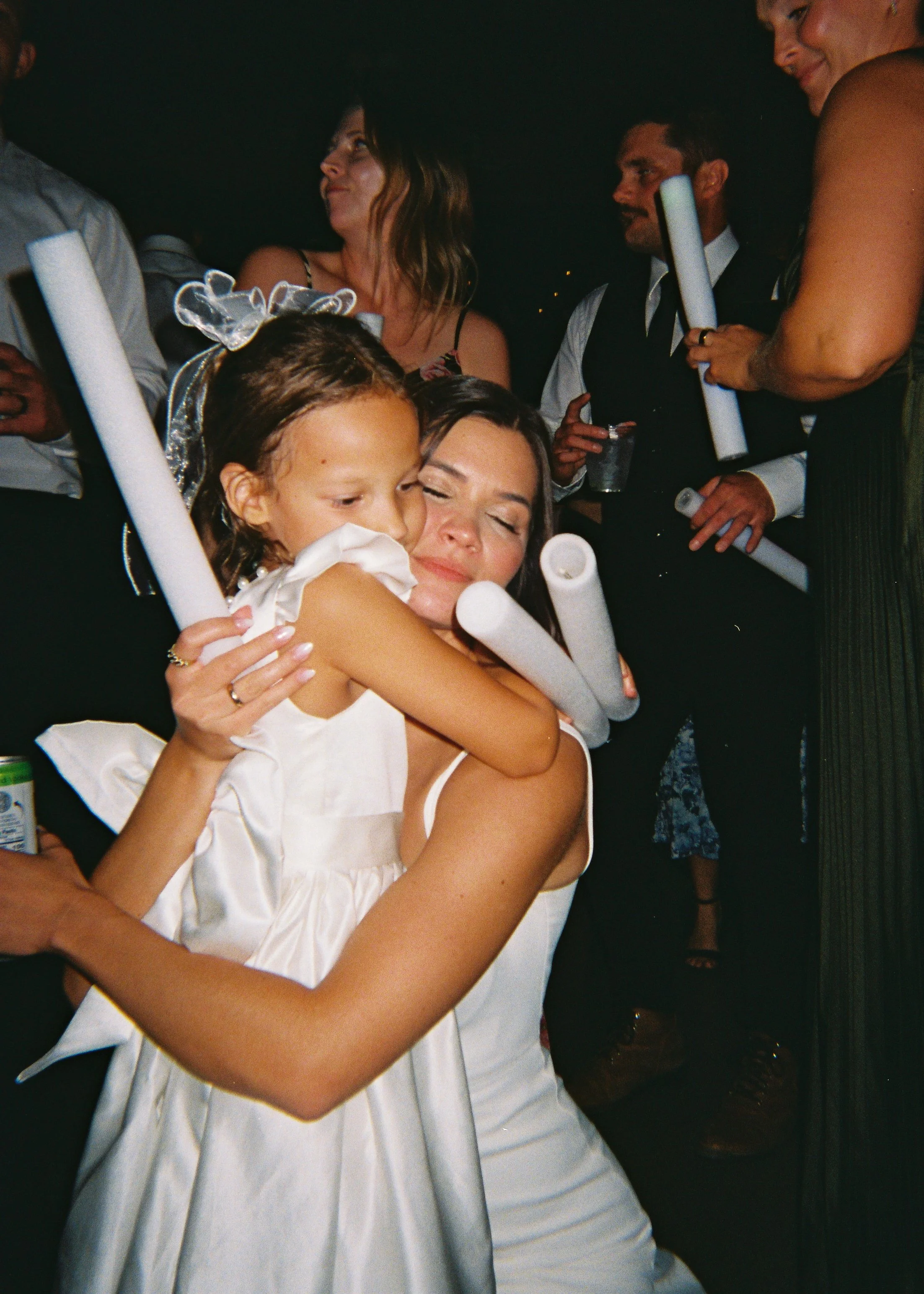 A group of people celebrating at a party or event, with a young girl hugging a woman, both smiling. The woman has her eyes closed, and the girl has curly hair and a bow in her hair. Other adults are in the background, holding papers and drinks.