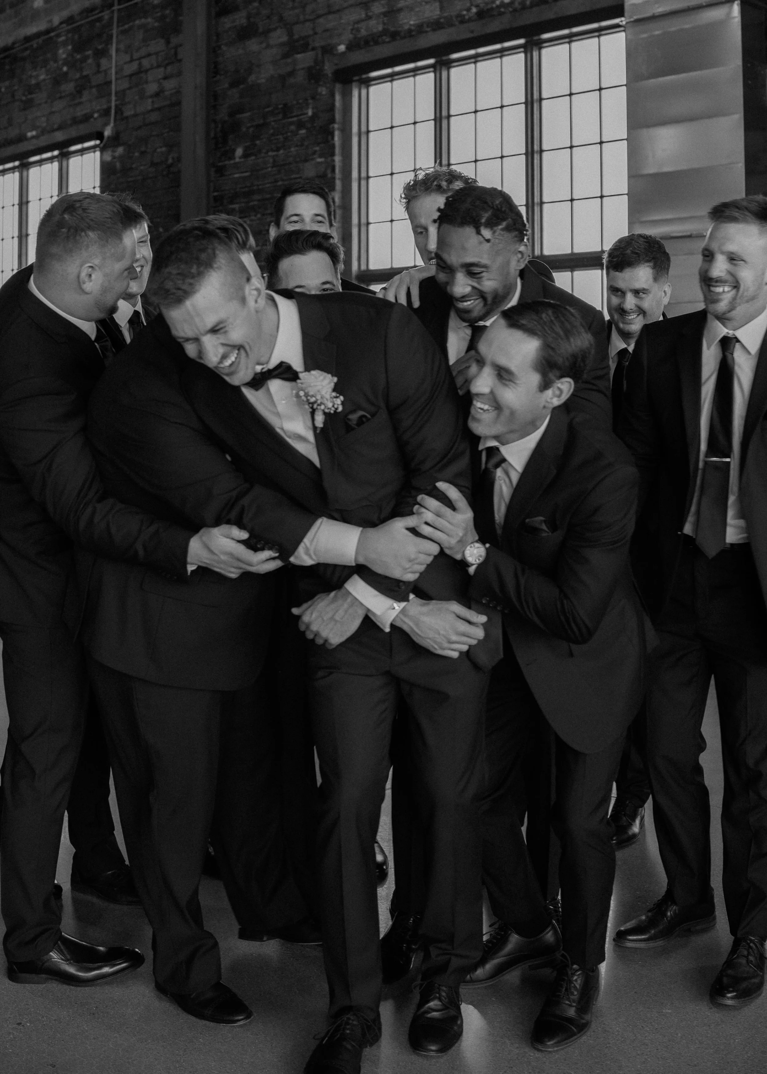 A group of men in suits laughing and hugging with one of them in the center leaning forward. They are standing indoors near large windowpanes with an industrial brick wall in the background.