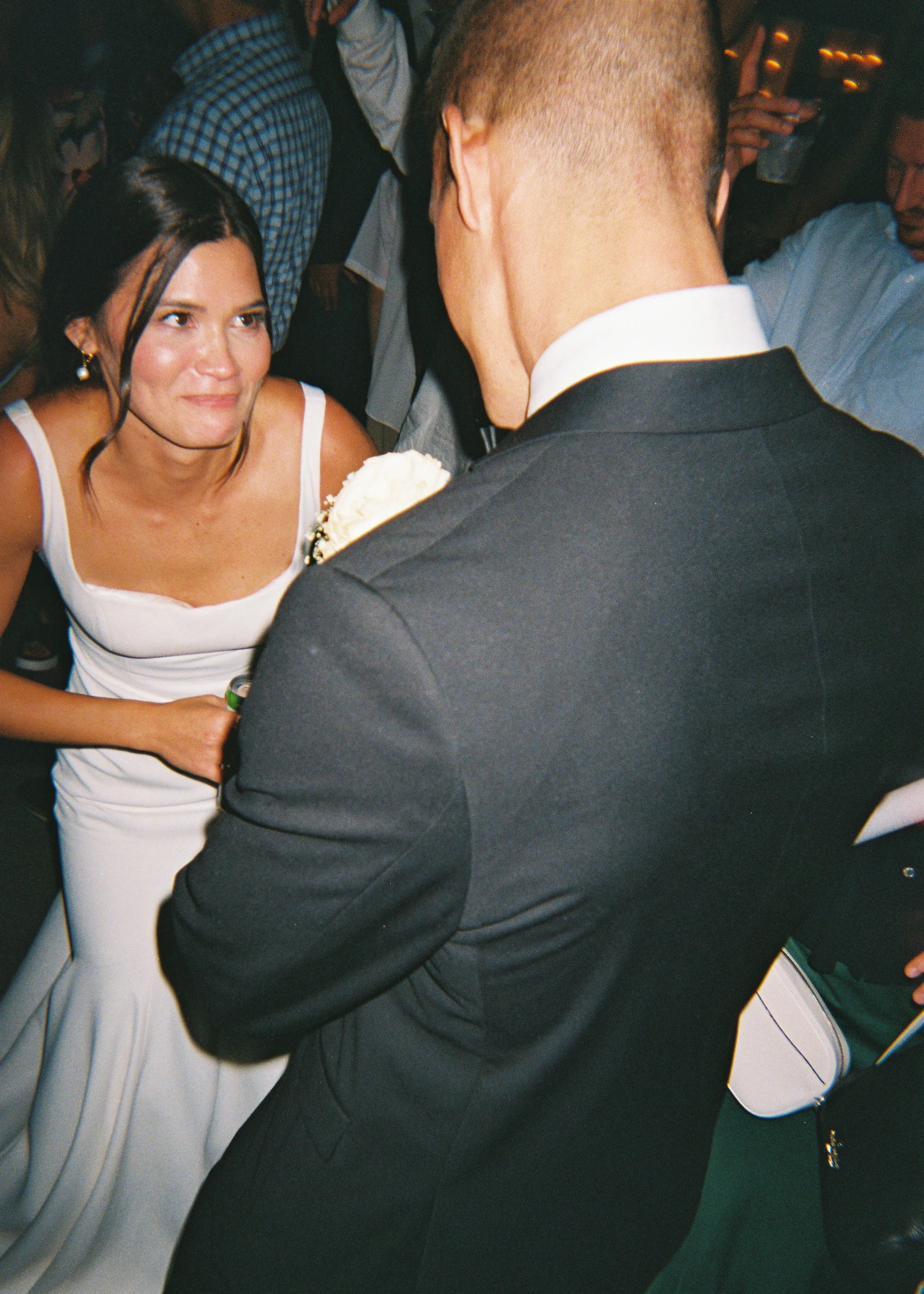 A woman in a white dress and a man in a black tuxedo with a white shirt are face to face at a dance or social event, with other people in the background.