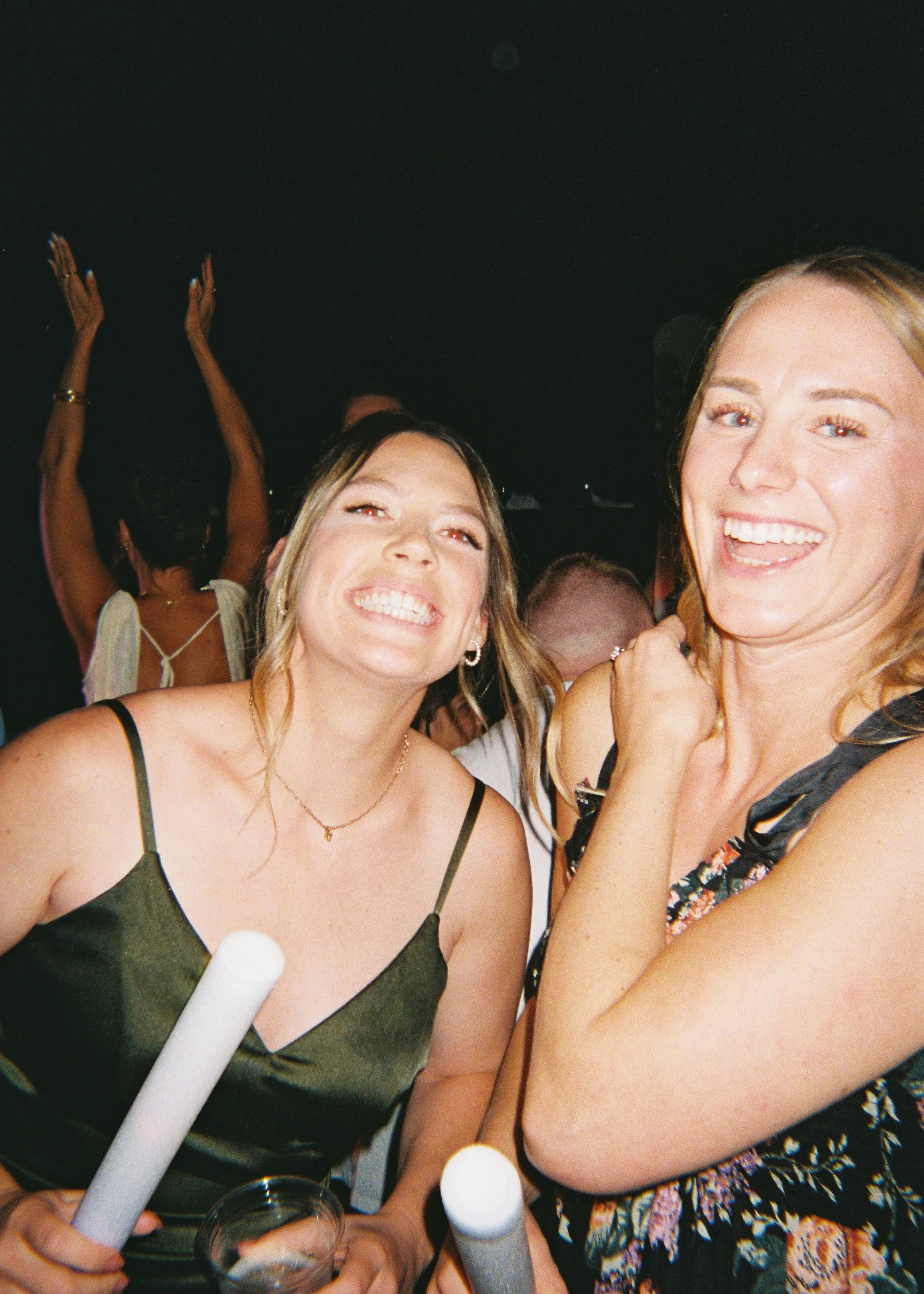 People at a celebration or party, smiling, with some holding rolled napkins and drinks, in a dimly lit environment.