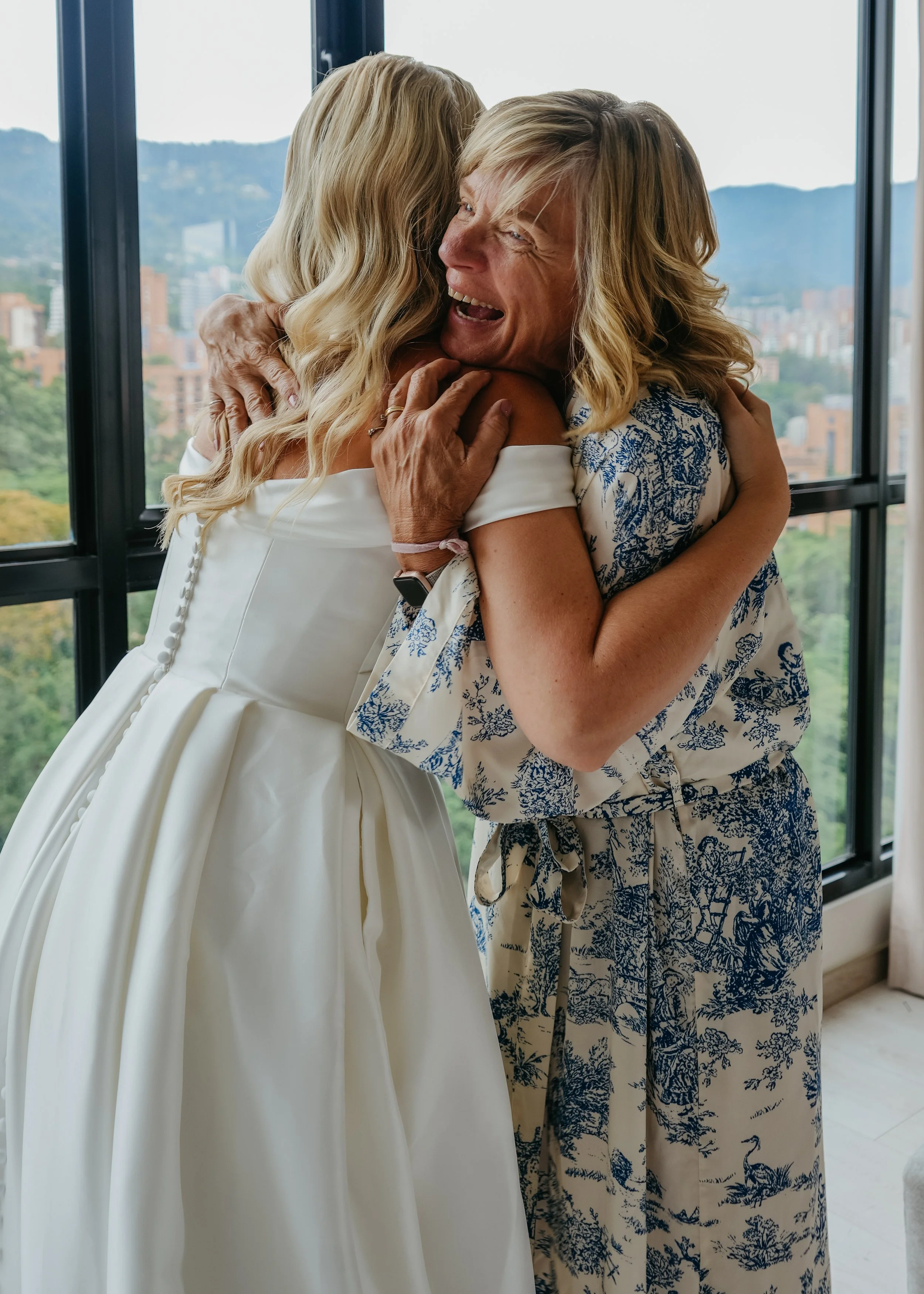Two women hugging each other joyfully in front of large windows with a cityscape and mountains in the background.