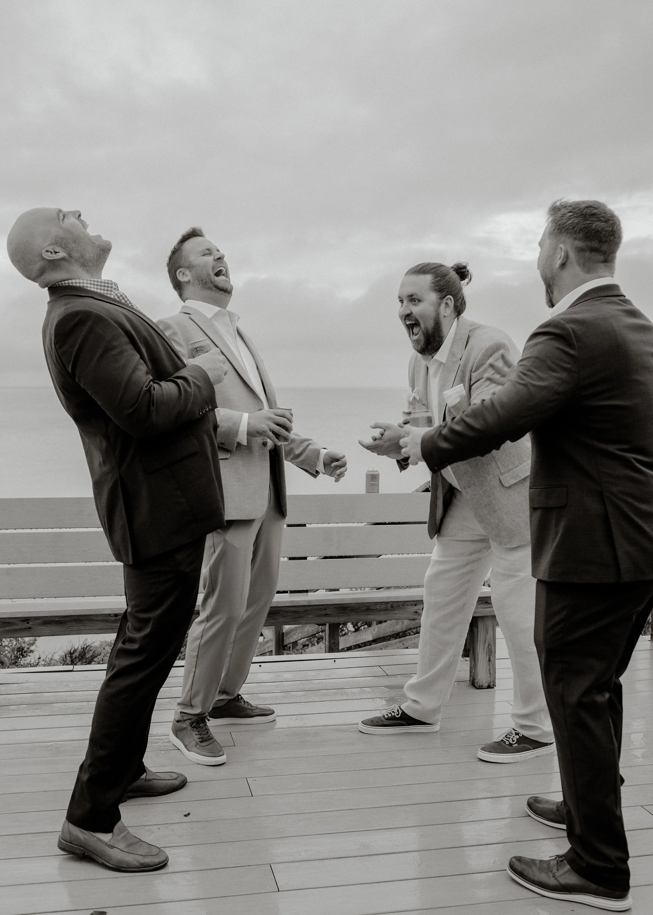 Four men in suits laughing and talking on a wooden outdoor deck with a cloudy sky in the background.