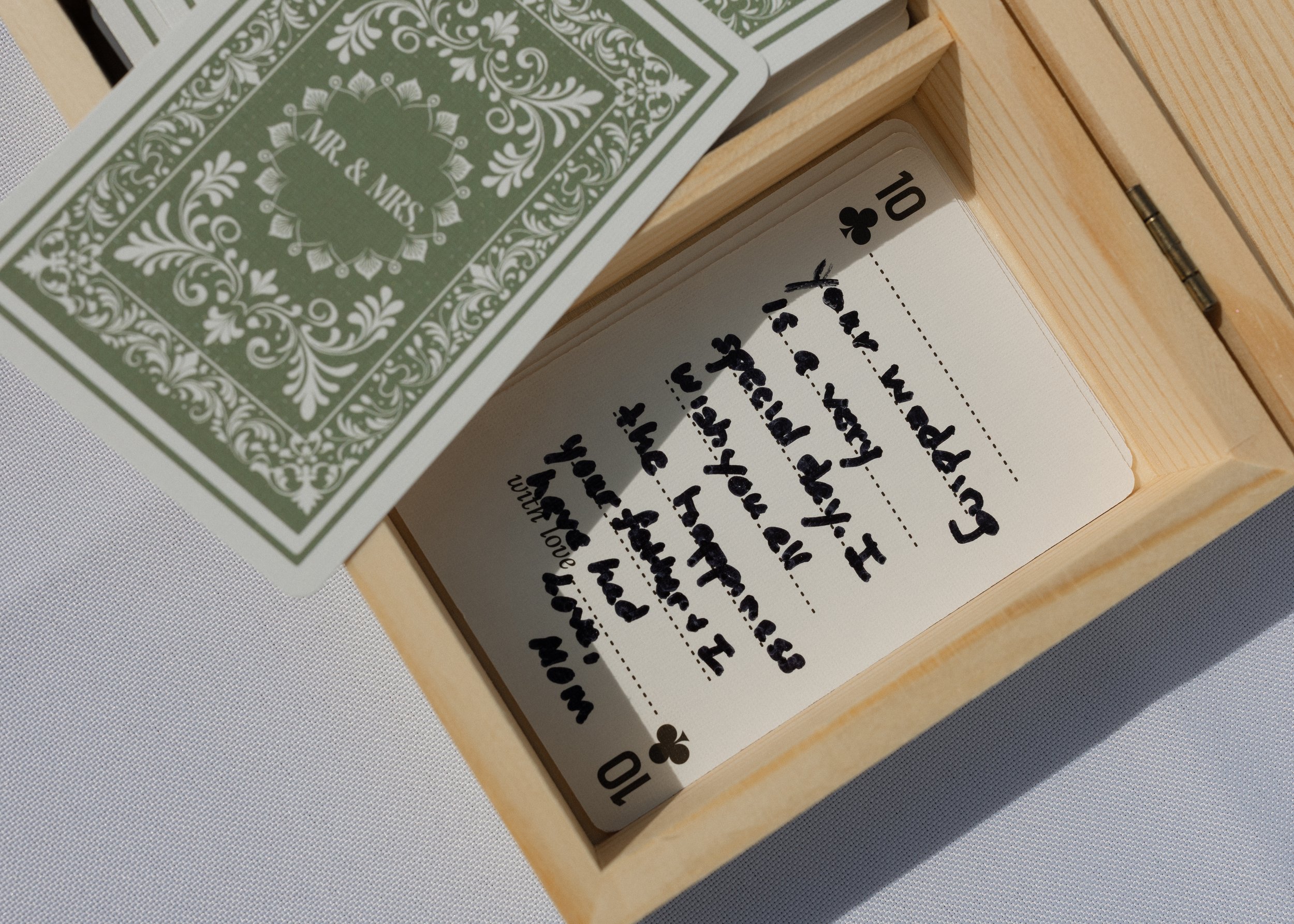 A deck of playing cards in a wooden box, with the top card being a 10 of Clubs, partially covering a handwritten note inside the box.
