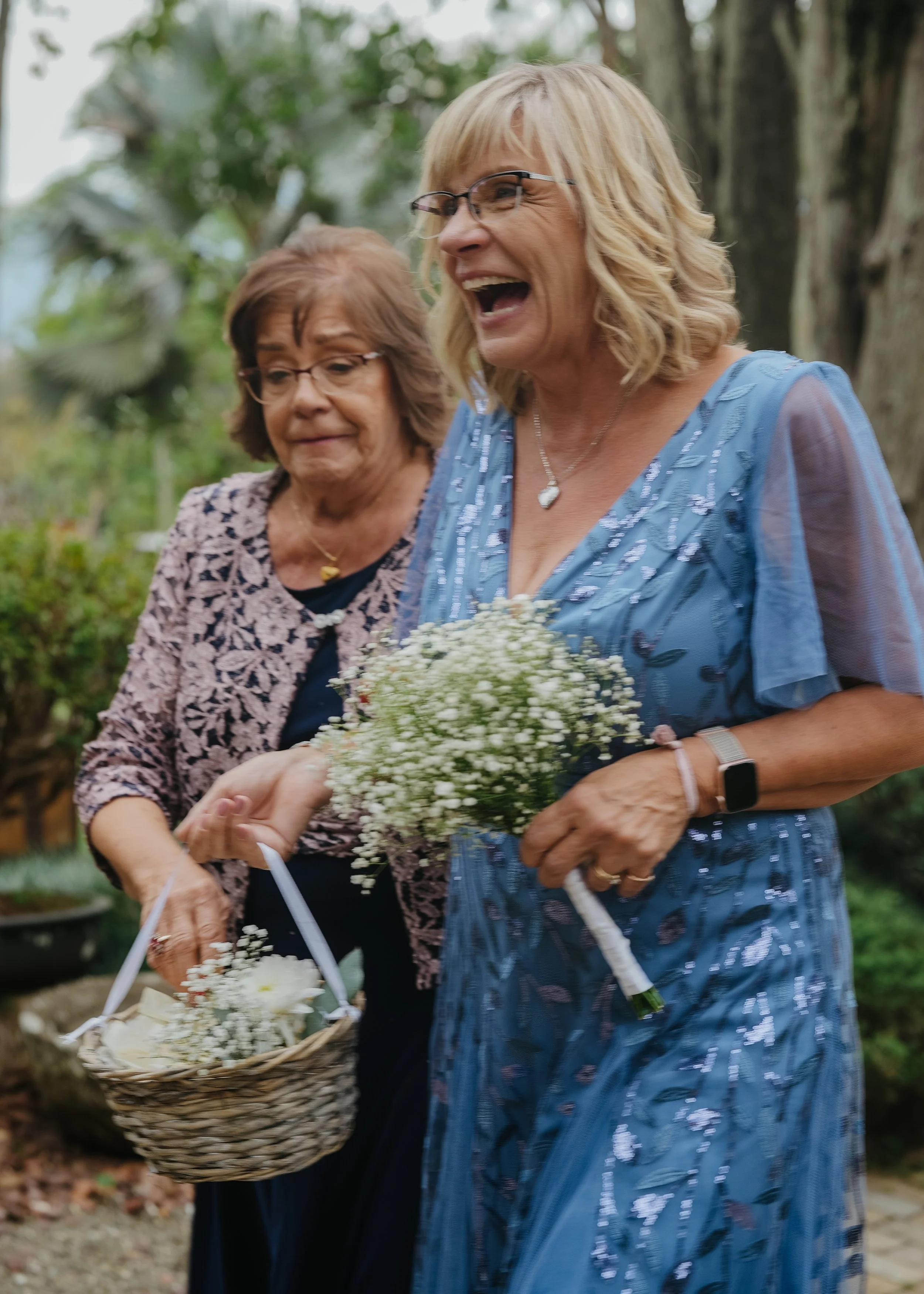 Two women, one with glasses and blonde hair wearing a blue dress, and the other with glasses and brown hair, are smiling and carrying baskets of white flowers outdoors surrounded by trees.