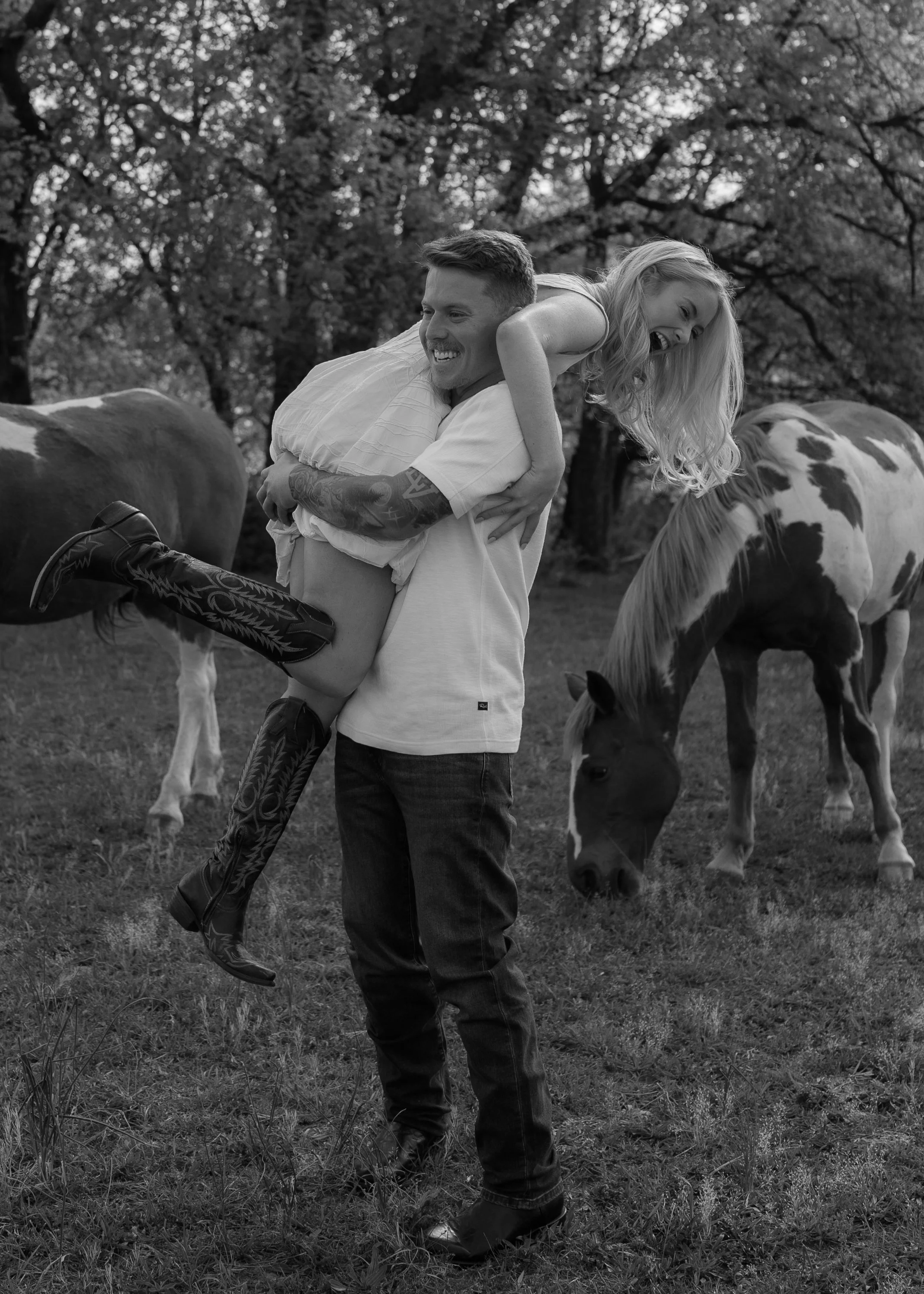 A man is giving a piggyback ride to a woman in a field with horses grazing and trees in the background.