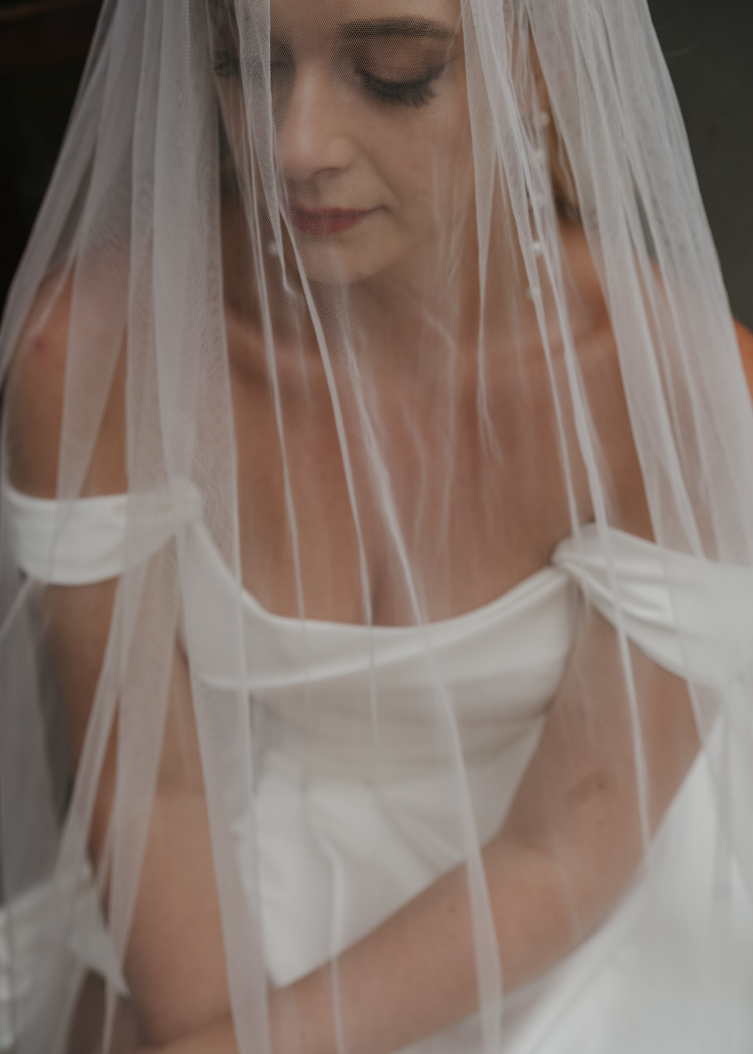 A woman with closed eyes wearing a soft veil over her face and shoulders, dressed in a white dress. The photo is taken through a textured glass, creating a distorted effect.