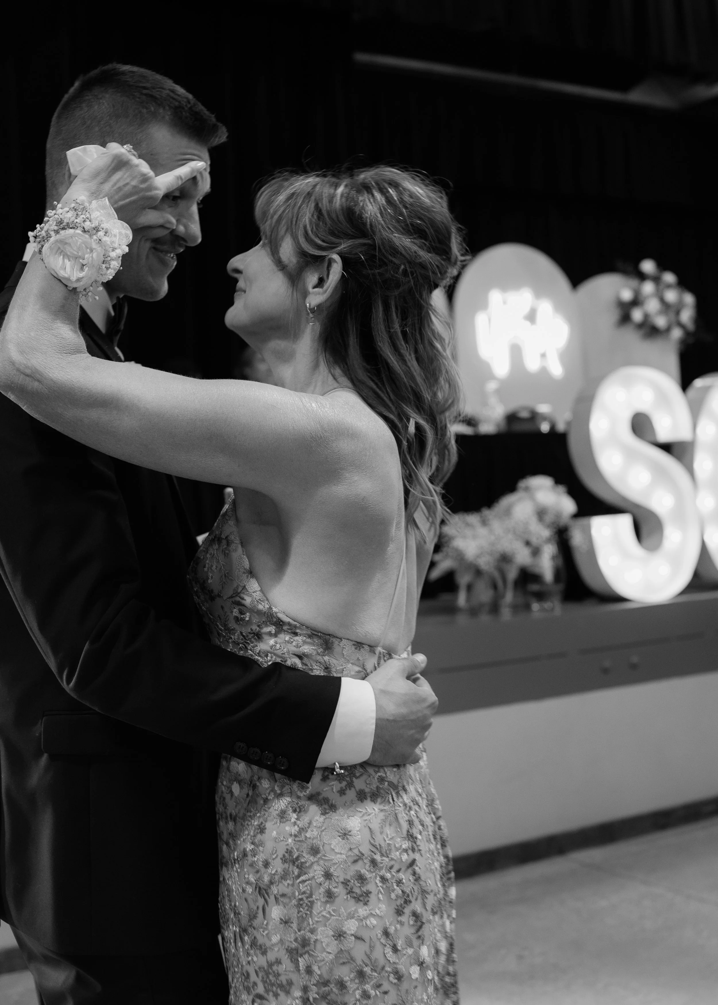 A young man and an older woman are dancing together, gazing into each other's eyes. The man is wearing a suit with a boutonniere, and the woman is wearing a floral dress. Their foreheads are touching, and she has her arms around his neck. The backgro