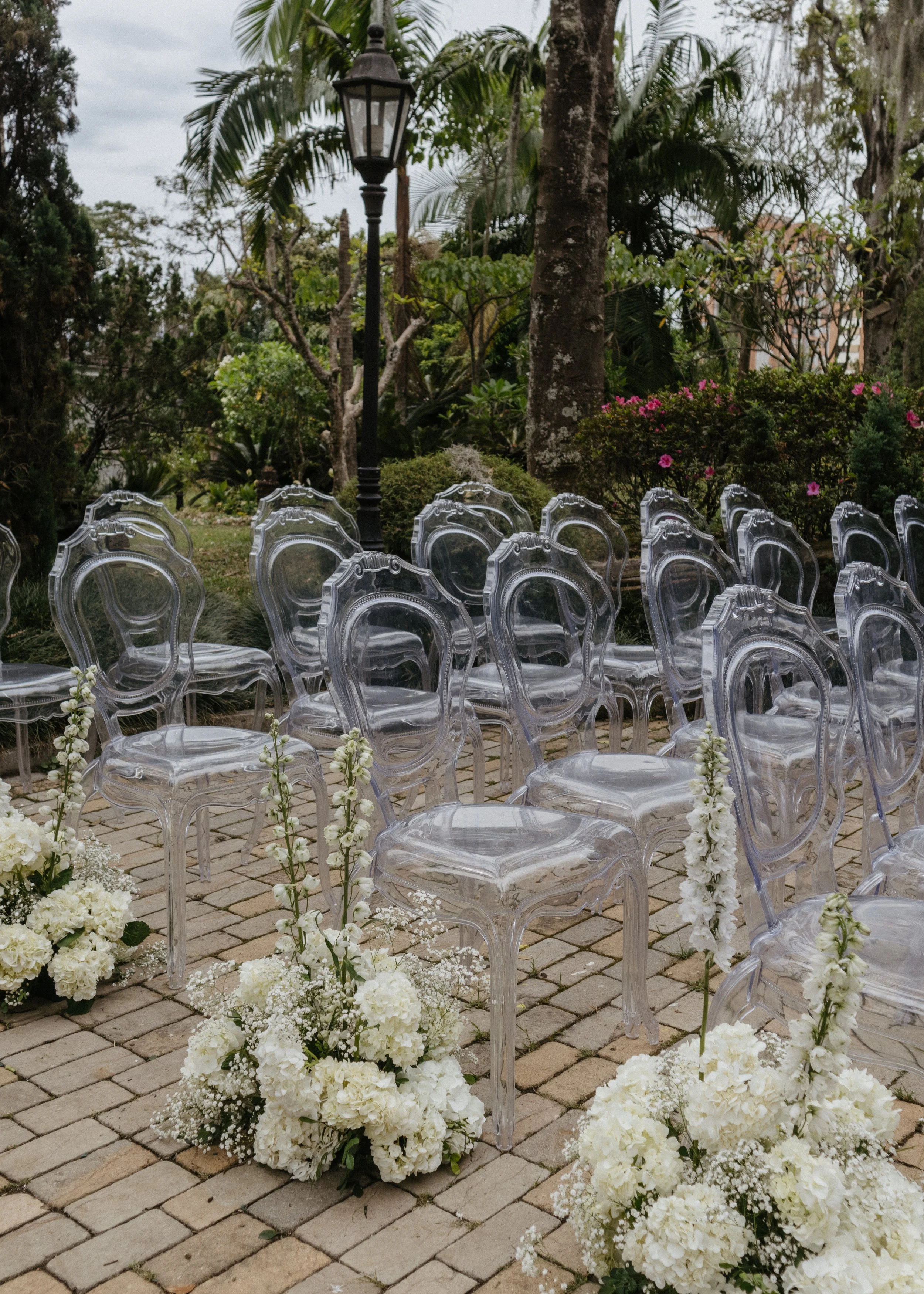 Outdoor wedding ceremony setup with transparent ghost chairs, white floral arrangements, and a black lamppost, surrounded by trees and greenery.