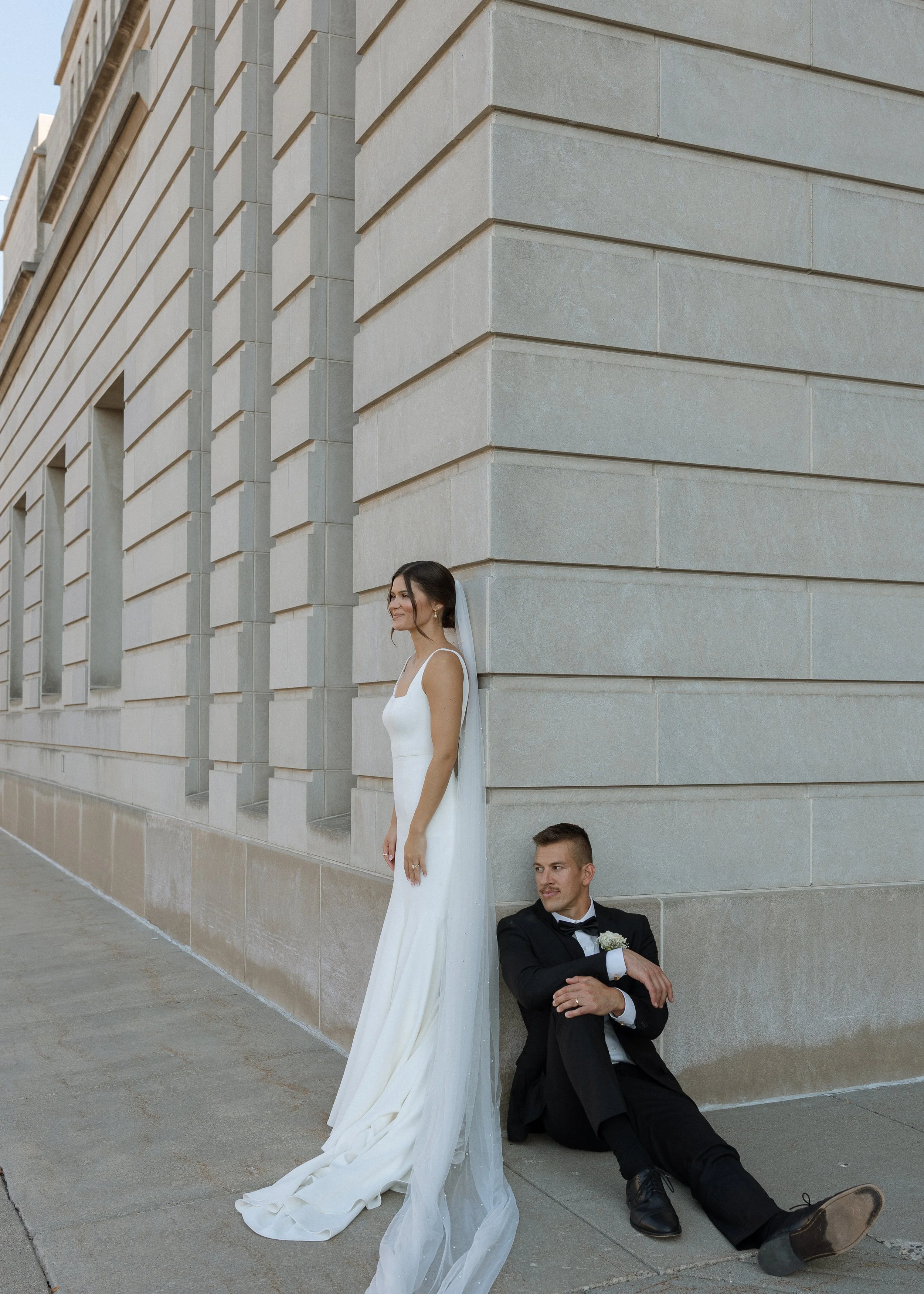 A bride in a white wedding gown with a long train stands outdoors against a building with large stone blocks, looking into the distance with a slight smile. A groom in a black tuxedo and bow tie is sitting on the ground, leaning against the building,