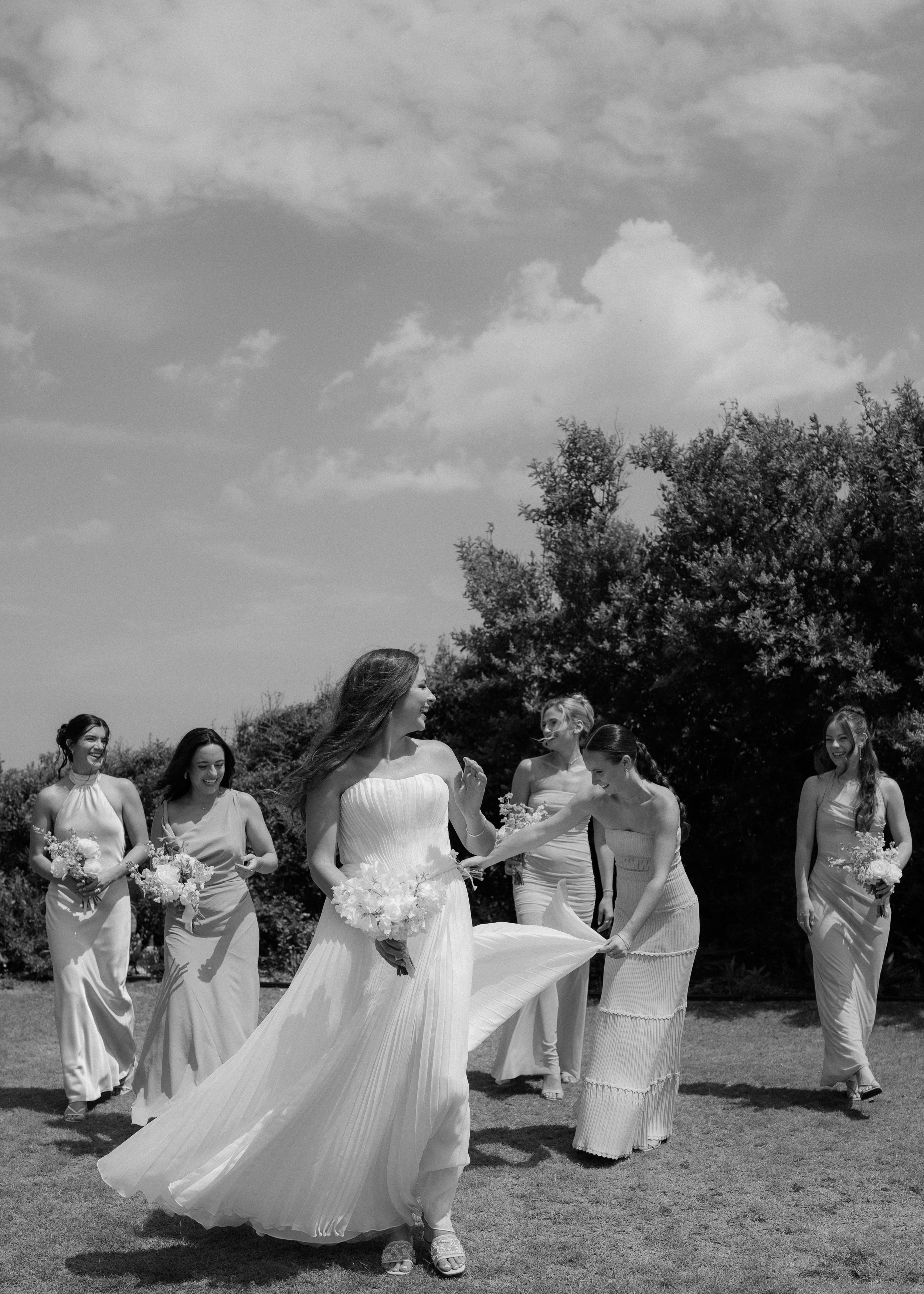 A bride surrounded by bridesmaids outdoors on a grassy field, holding bouquets, under a partly cloudy sky.