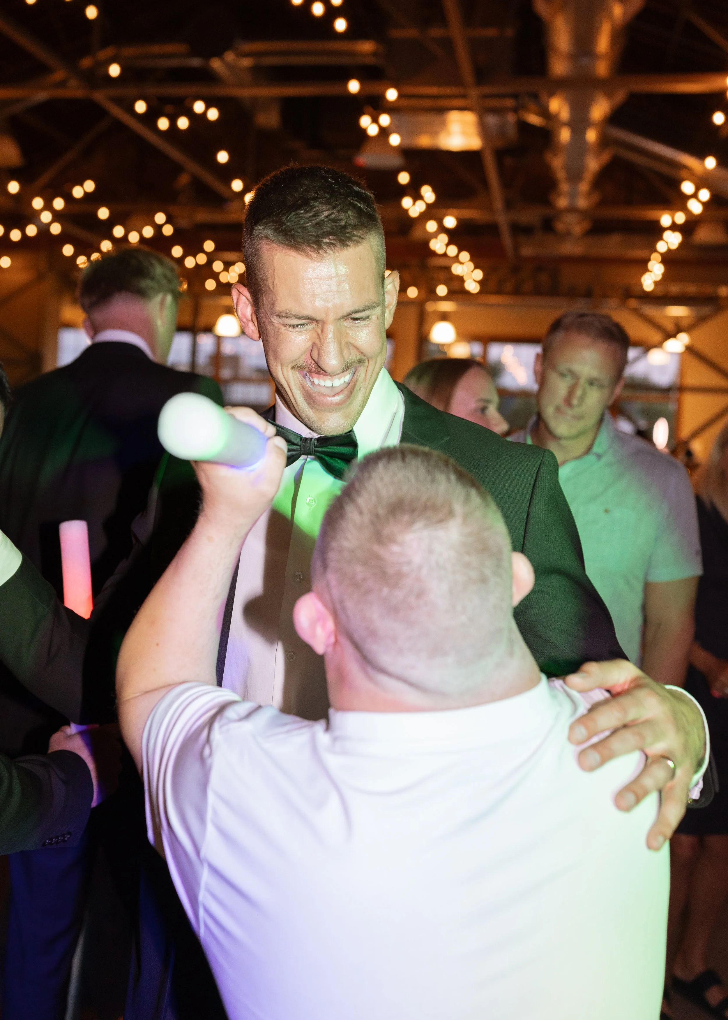 A group of men dressed in formal attire, including tuxedos, dance and celebrate at a party or wedding reception with warm lighting and string lights hanging from the ceiling.