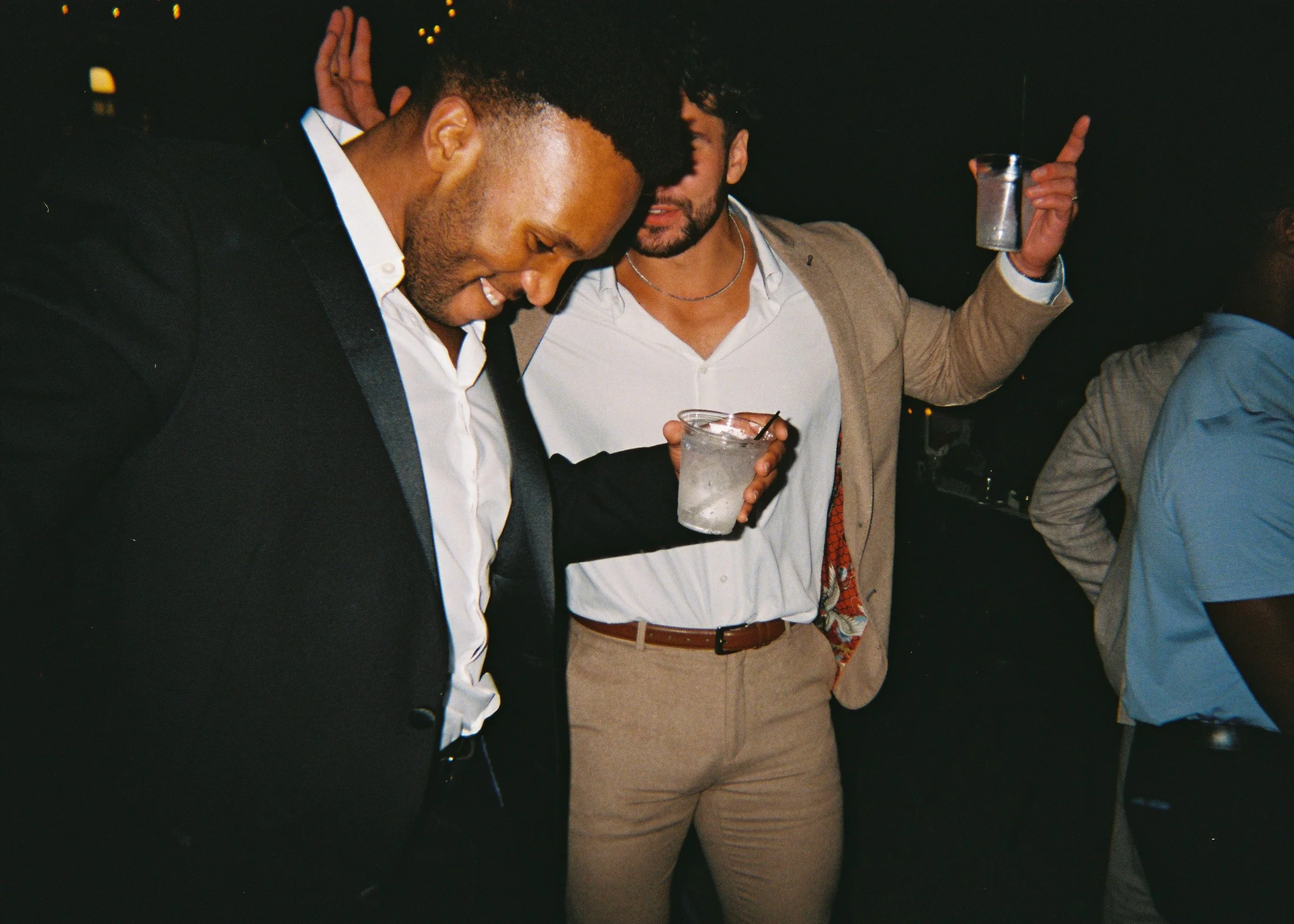 Two men at a social gathering are smiling and talking, one holding a drink, the other raising an arm with a drink in hand, with a dark background and blurry lights.