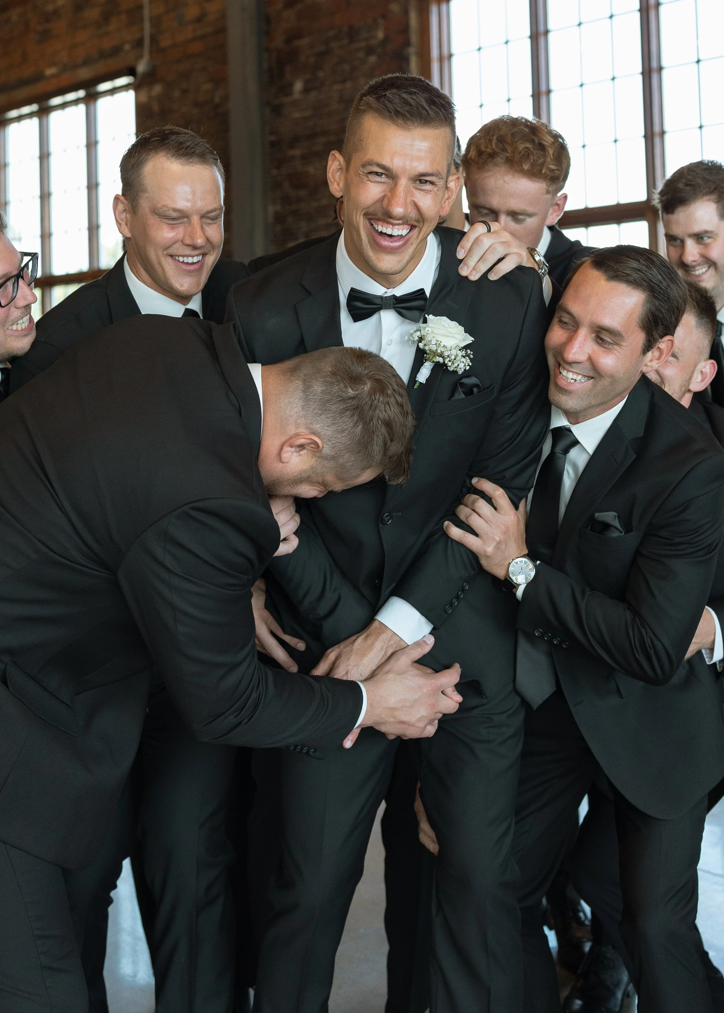 Group of men dressed in black tuxedos, some wearing bow ties, participating in a playful activity or game indoors with a brick wall and large windows in the background.