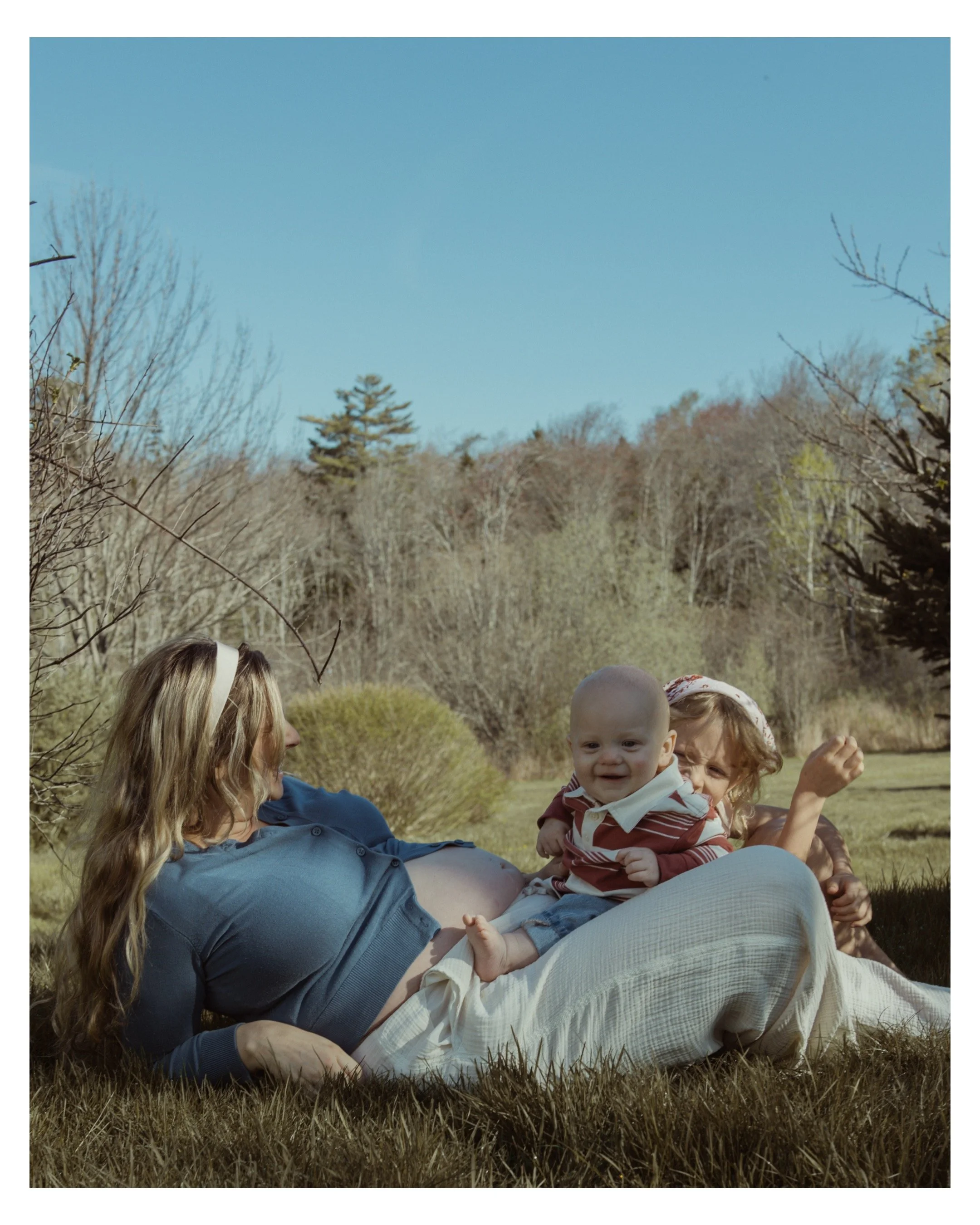 A family enjoying a sunny day outdoors on a grassy lawn, with trees and blue sky in the background. A woman with long blonde hair is lying on her back, holding a smiling baby in her lap, surrounded by other children and adults.