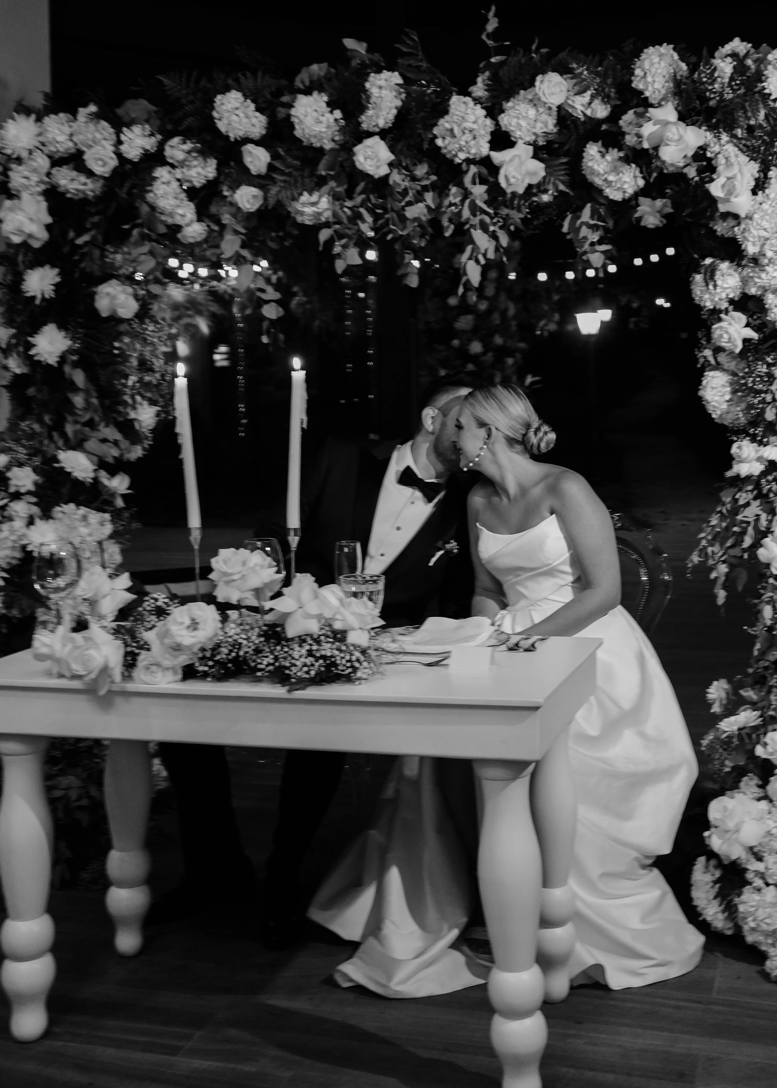 A bride and groom share a kiss at their wedding reception, seated at a table decorated with flowers, candles, and glasses, with a floral arch behind them.