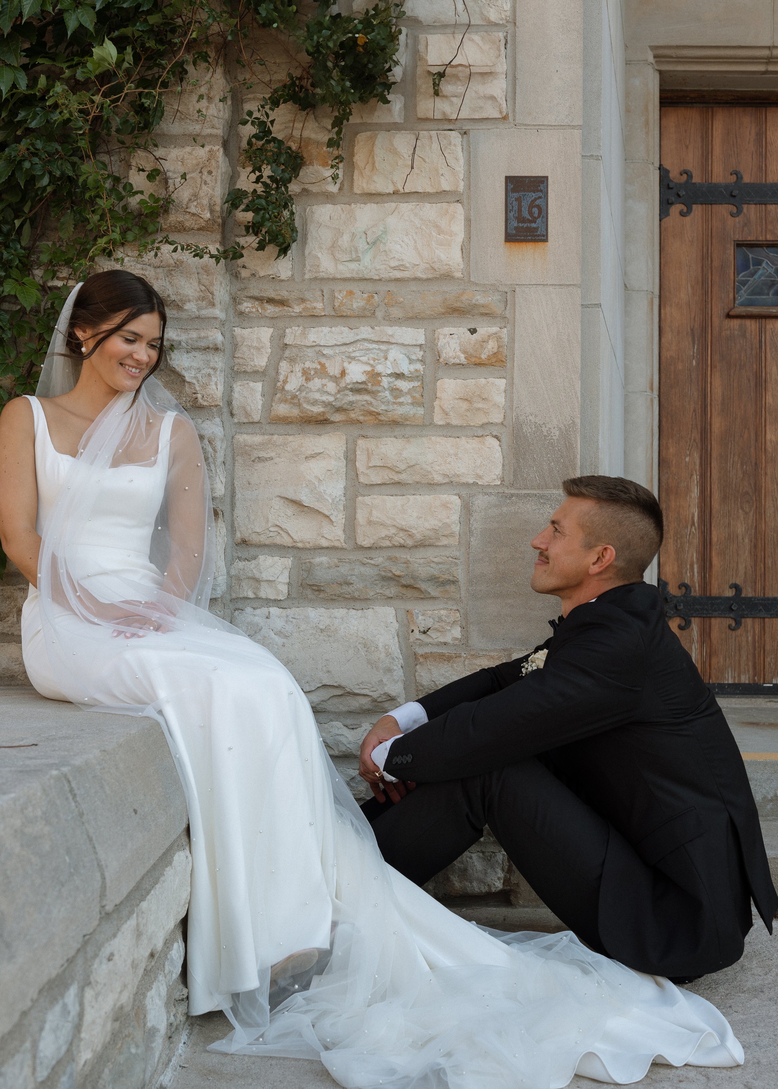 A bride in a white wedding dress and veil smiling at a groom in a black suit sitting on stairs outside a stone building with a wooden door and street number 16.