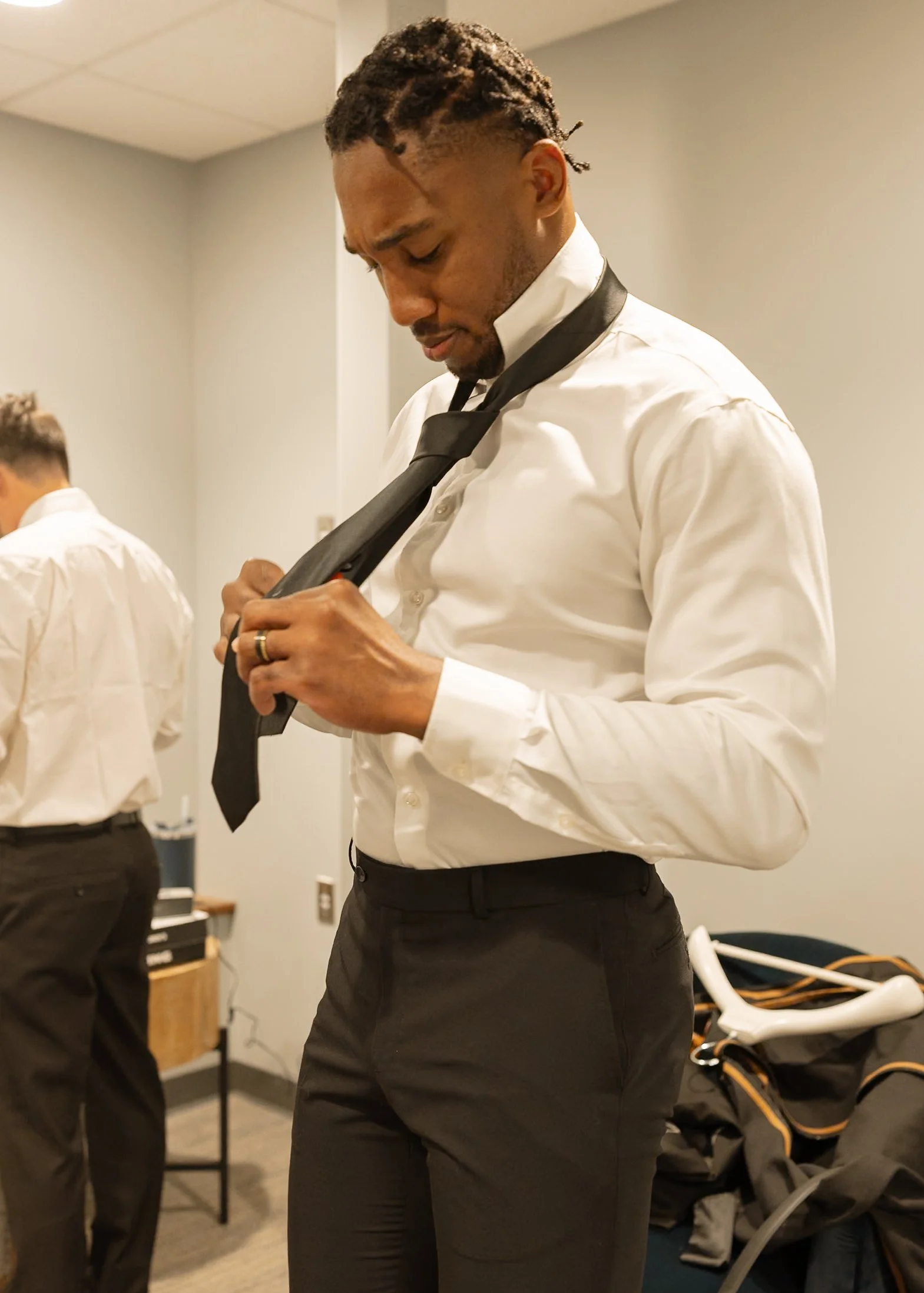 A man in dress pants and a white dress shirt adjusting his black tie in a room with gray walls, with another man and various items visible in the background.