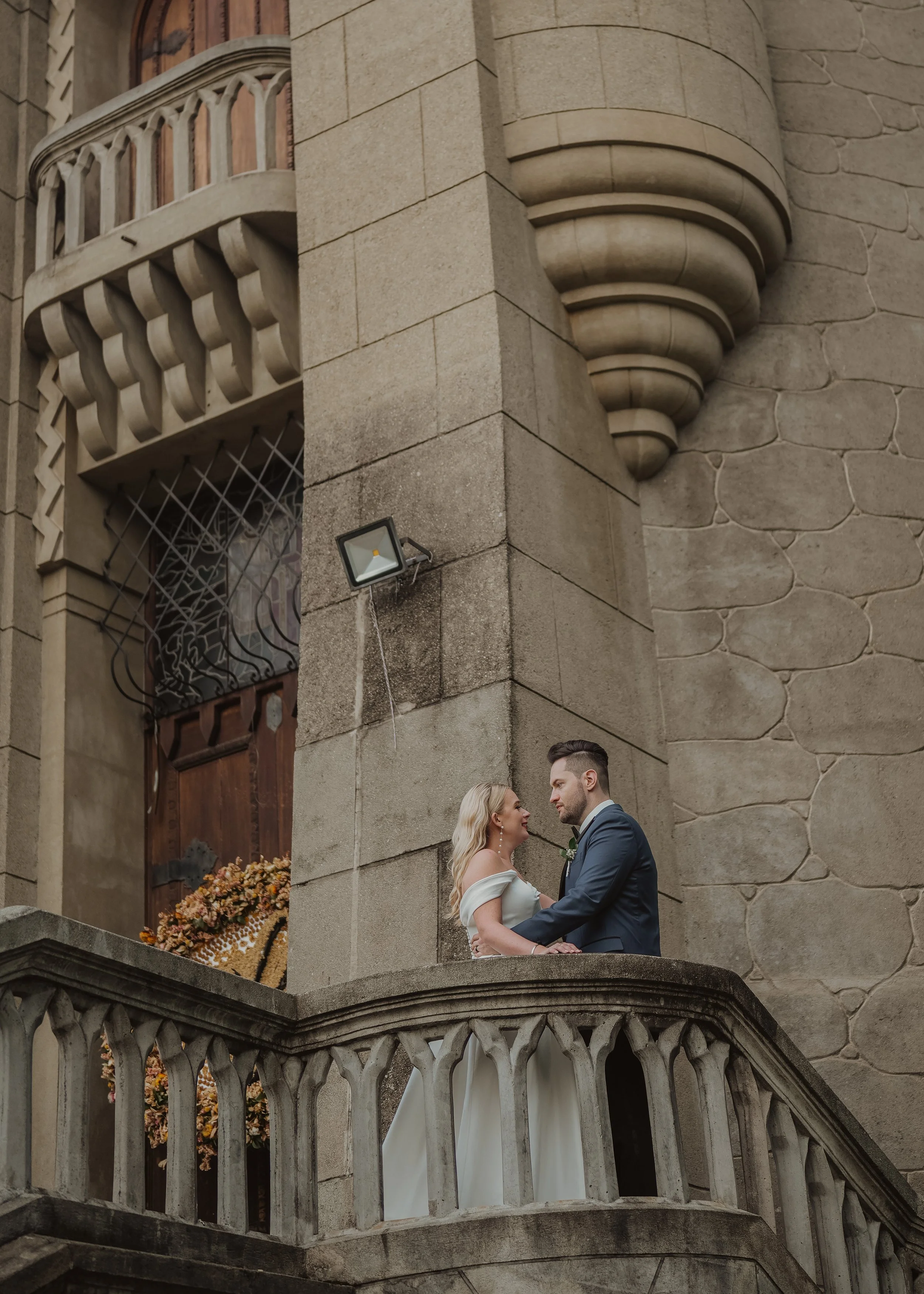 A couple dressed in wedding attire standing on a balcony of a historic stone building with decorative architectural details, holding hands and gazing into each other's eyes.