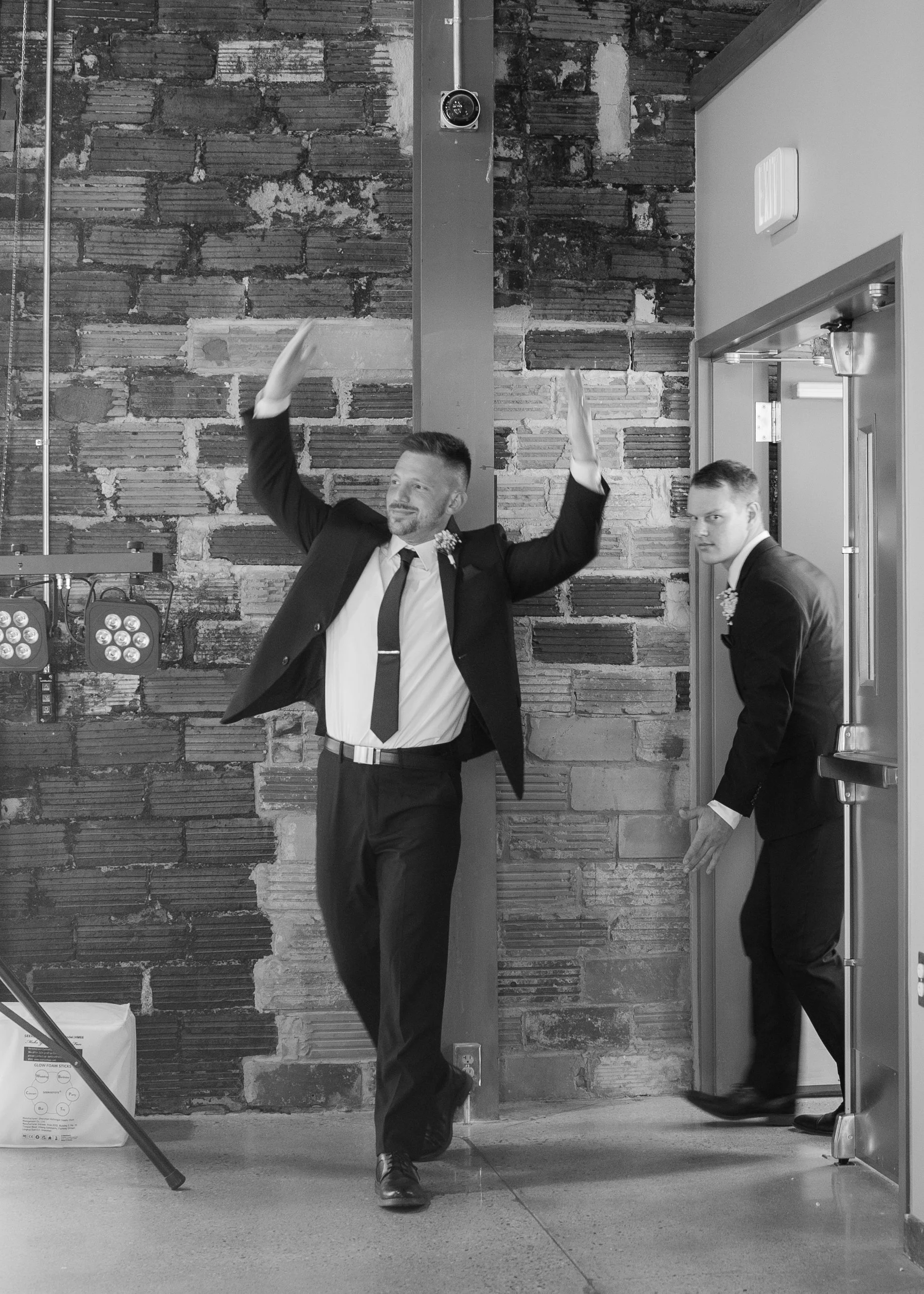Two men in suits, one with a beard, standing and posing in an industrial-style room with a brick wall background. One man is leaning casually, and the other is looking at the camera near an elevator door.