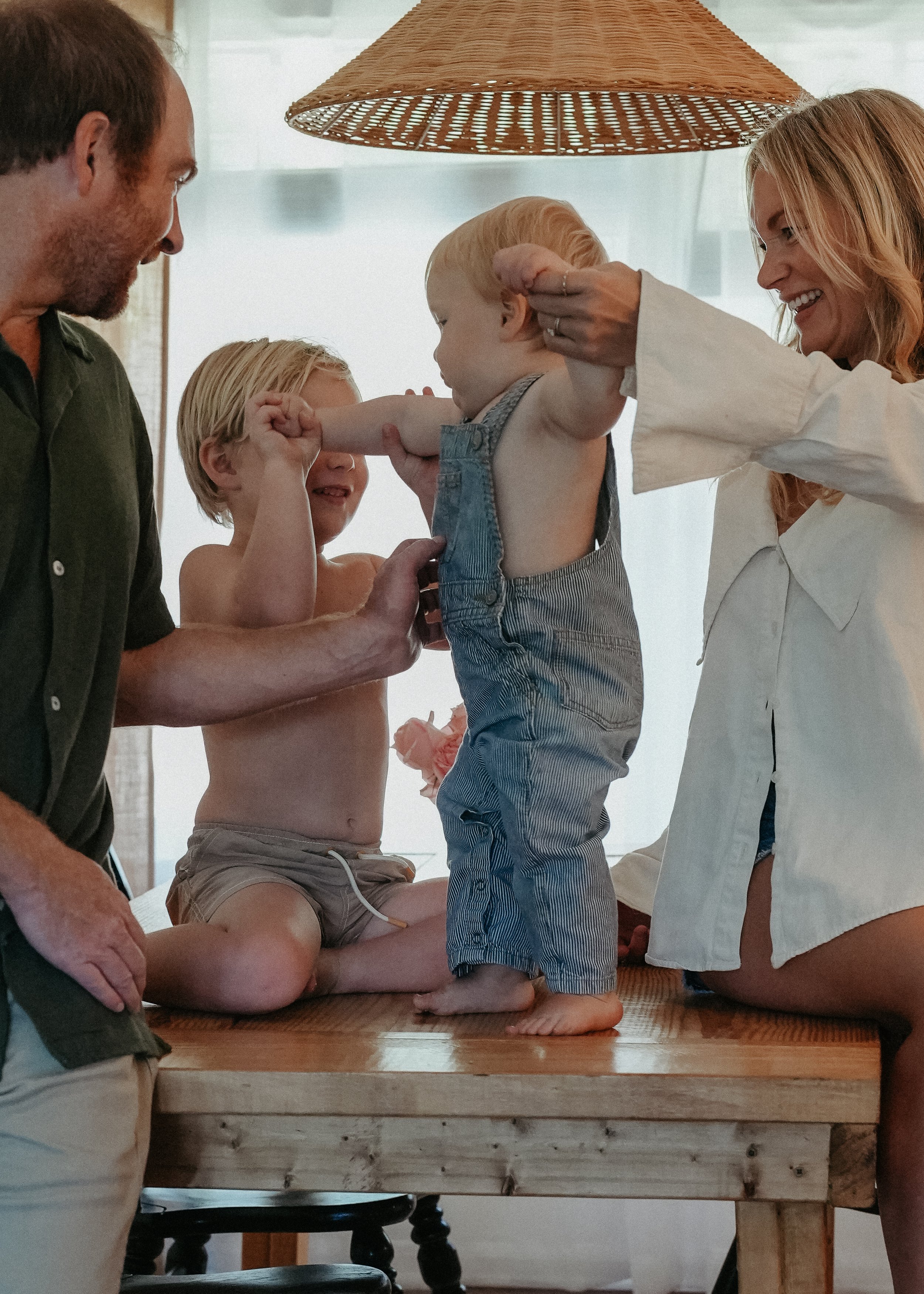 A family of four, including a man, a woman, and two young boys with blonde hair, gathered around a wooden table. The woman is smiling while helping a young boy stand and pose with his arm flexed. The father, who is shirtless, and the other child are watching and smiling in a cozy, well-lit room.