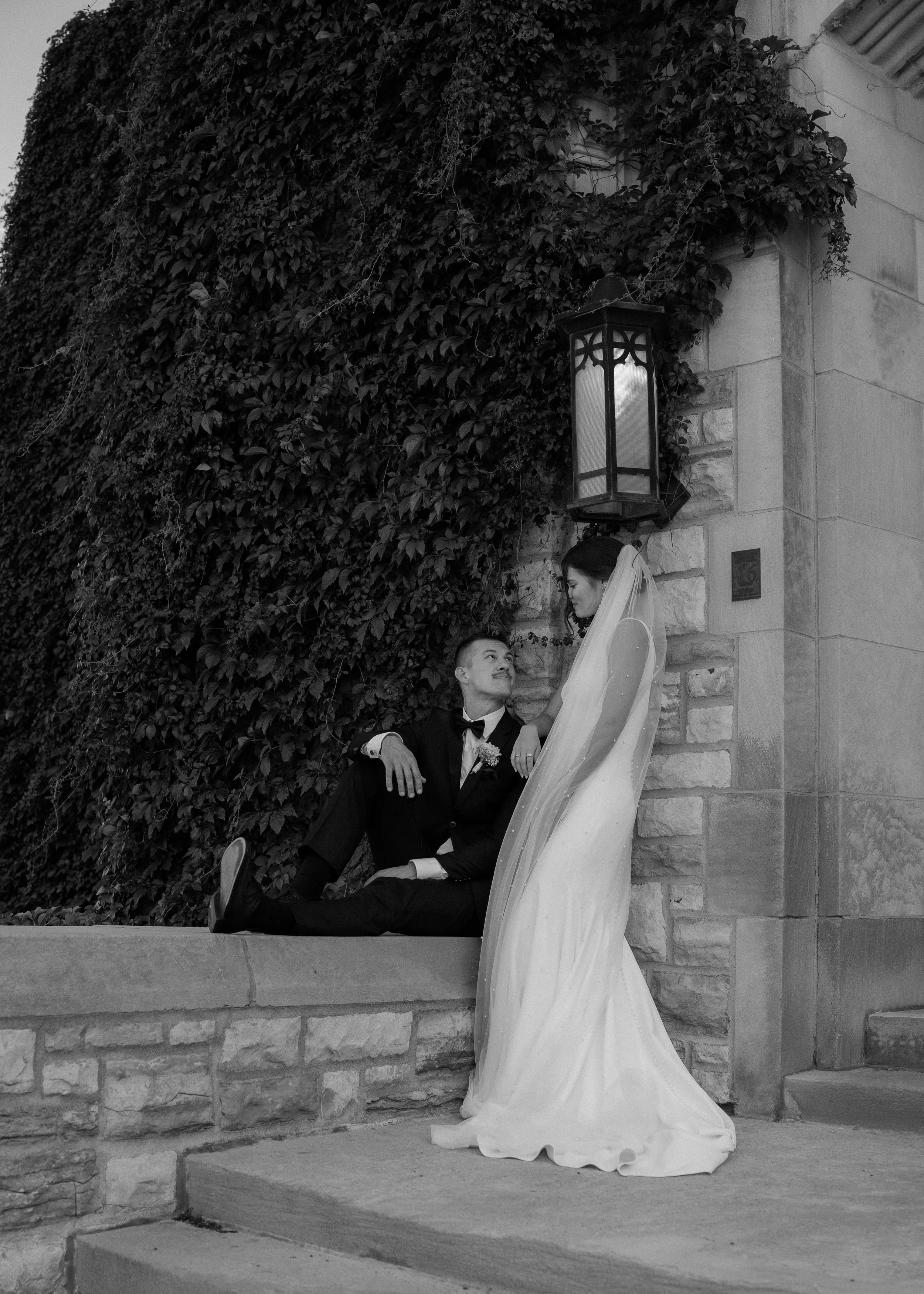 A black-and-white photo of a newlywed couple posing against a stone wall covered with ivy. The groom, dressed in a tuxedo, is sitting on the steps, looking up at his bride. The bride, wearing a wedding gown and veil, is standing slightly above him, gazing down with a gentle expression.