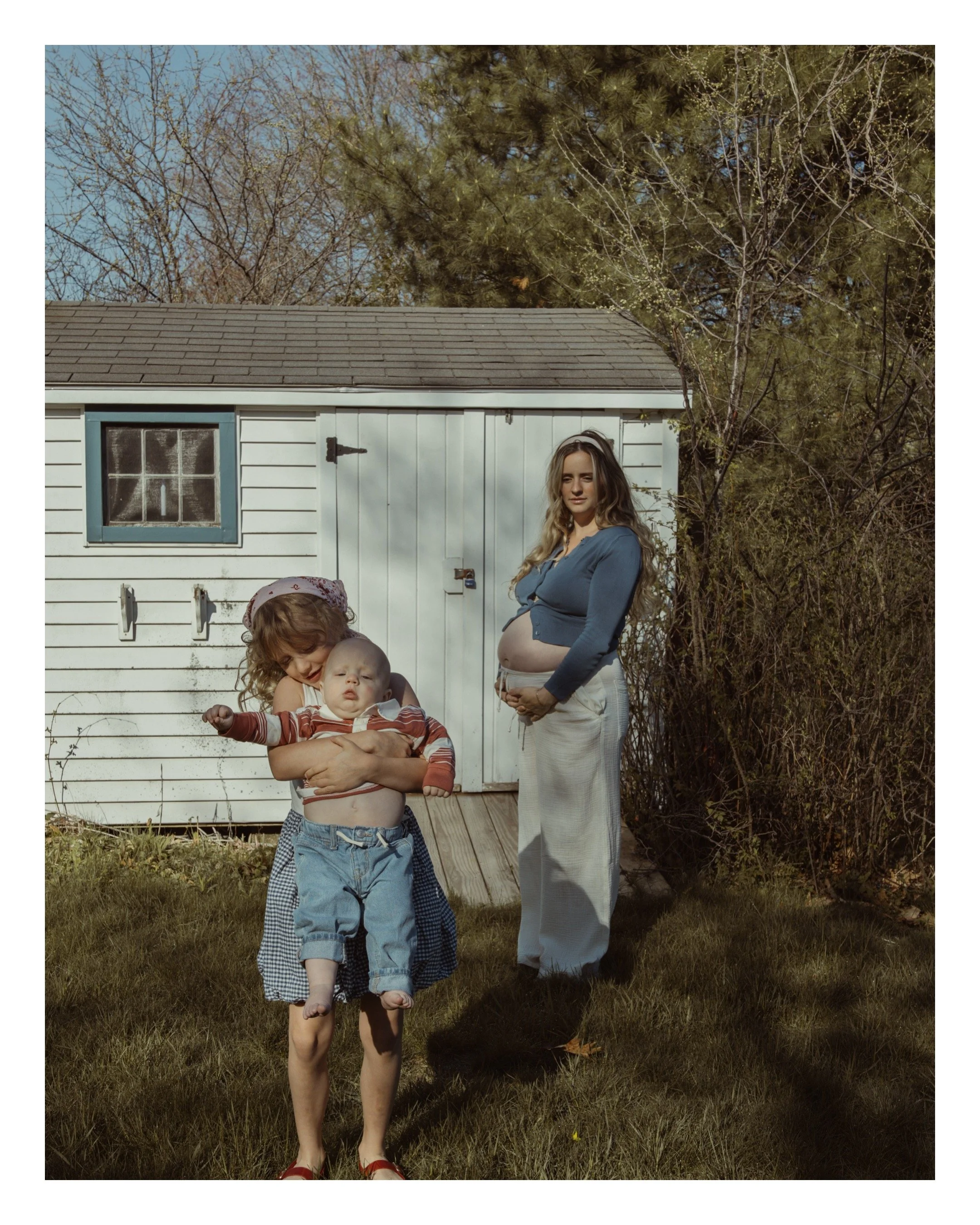 Young girl holding a baby with shared ultrasound pregnancy belly, woman with pregnant belly and crossed arms standing outside near a white shed and trees on sunny day.