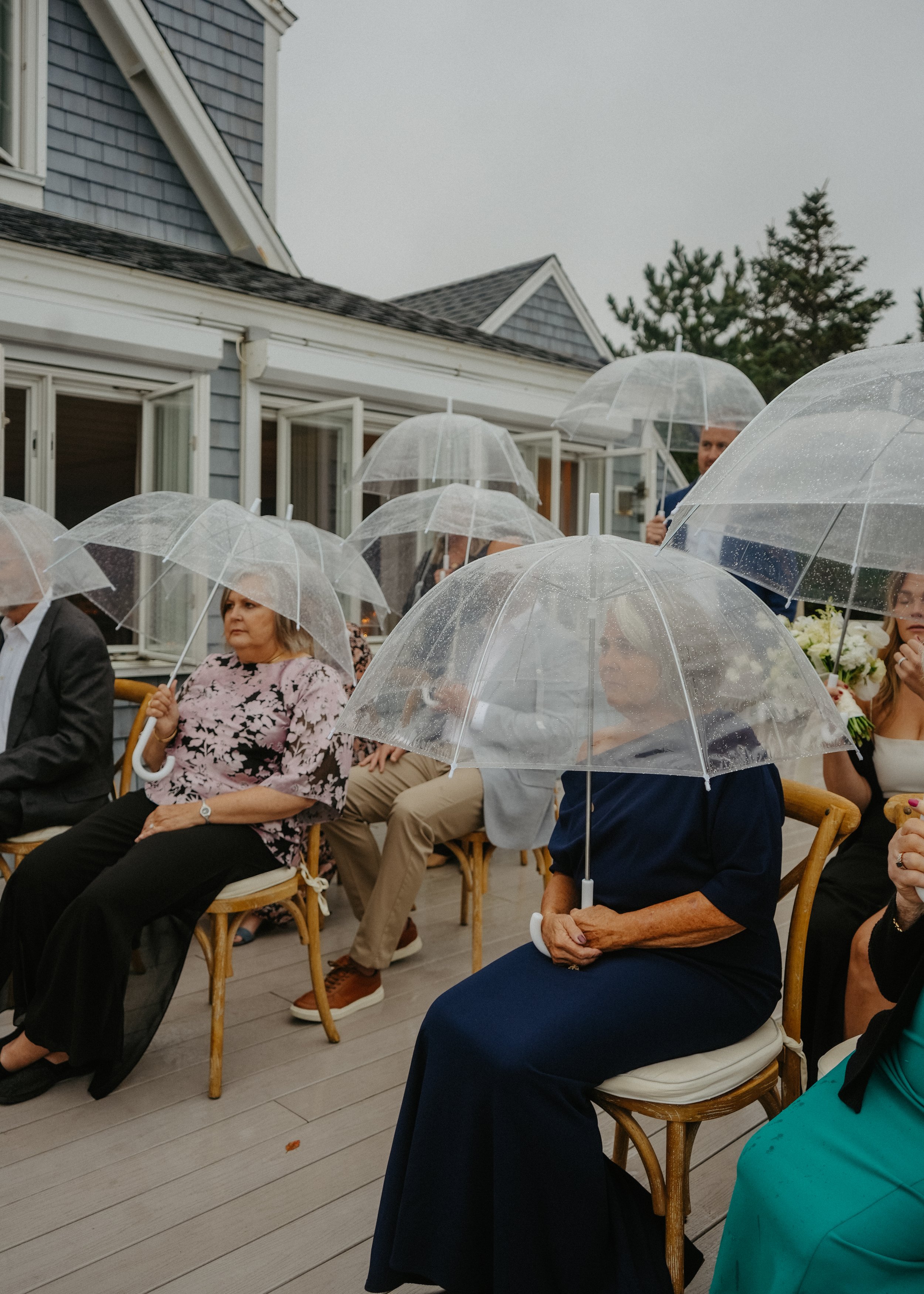 People sitting outdoors under umbrellas on a cloudy day, attending a gathering or ceremony in a backyard with a house in the background.