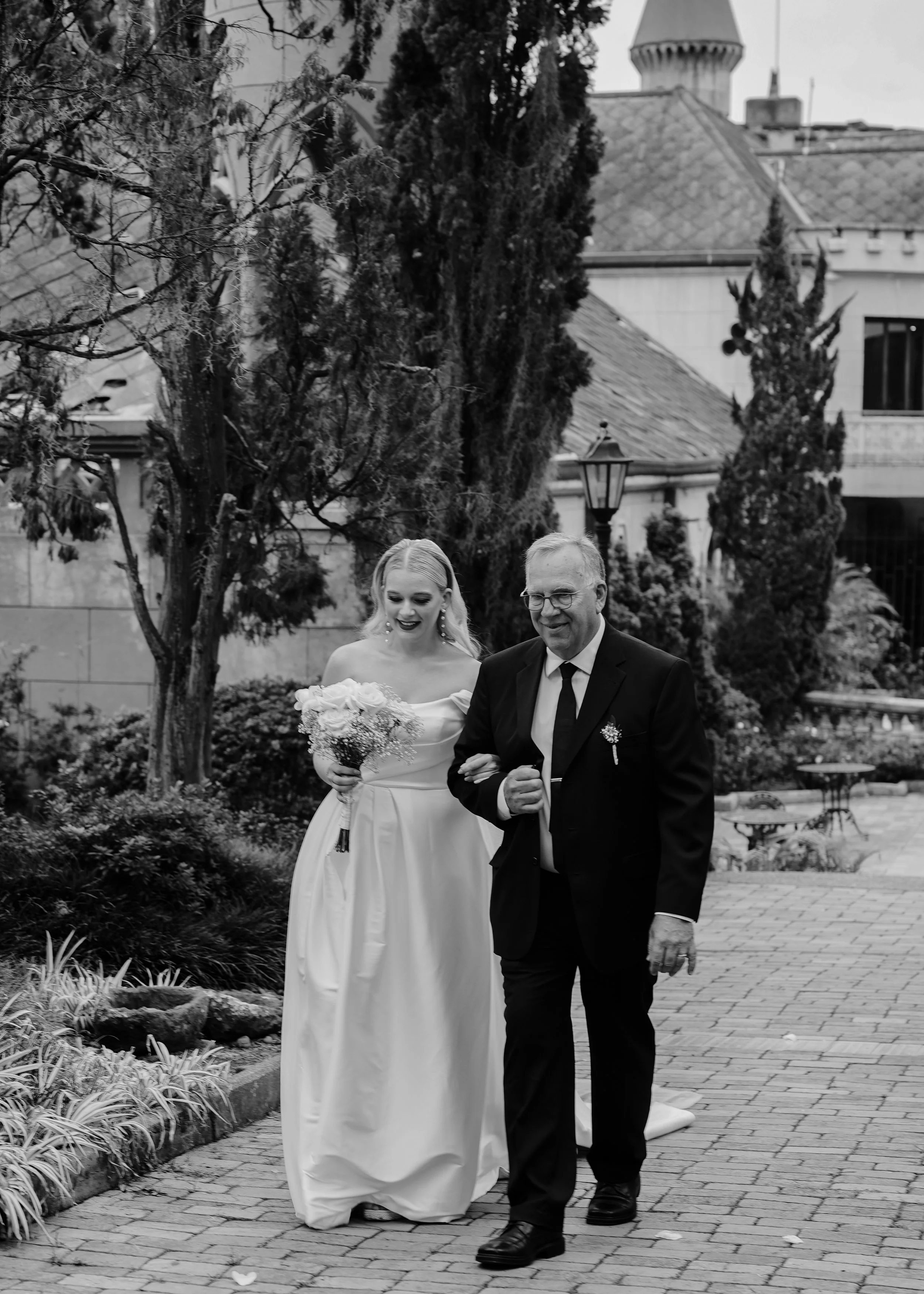 A bride and an older man walking arm in arm outdoors on a brick pathway, with trees and a building in the background, during a wedding celebration.