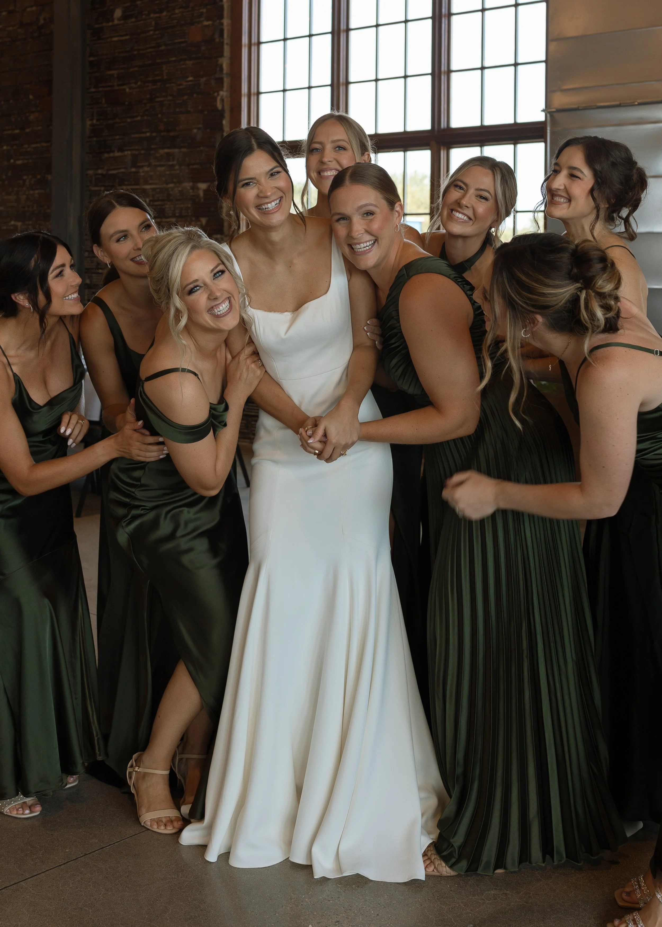 A group of nine women, including a bride in a white gown, smiling and hugging each other in a rustic indoor setting with large windows.